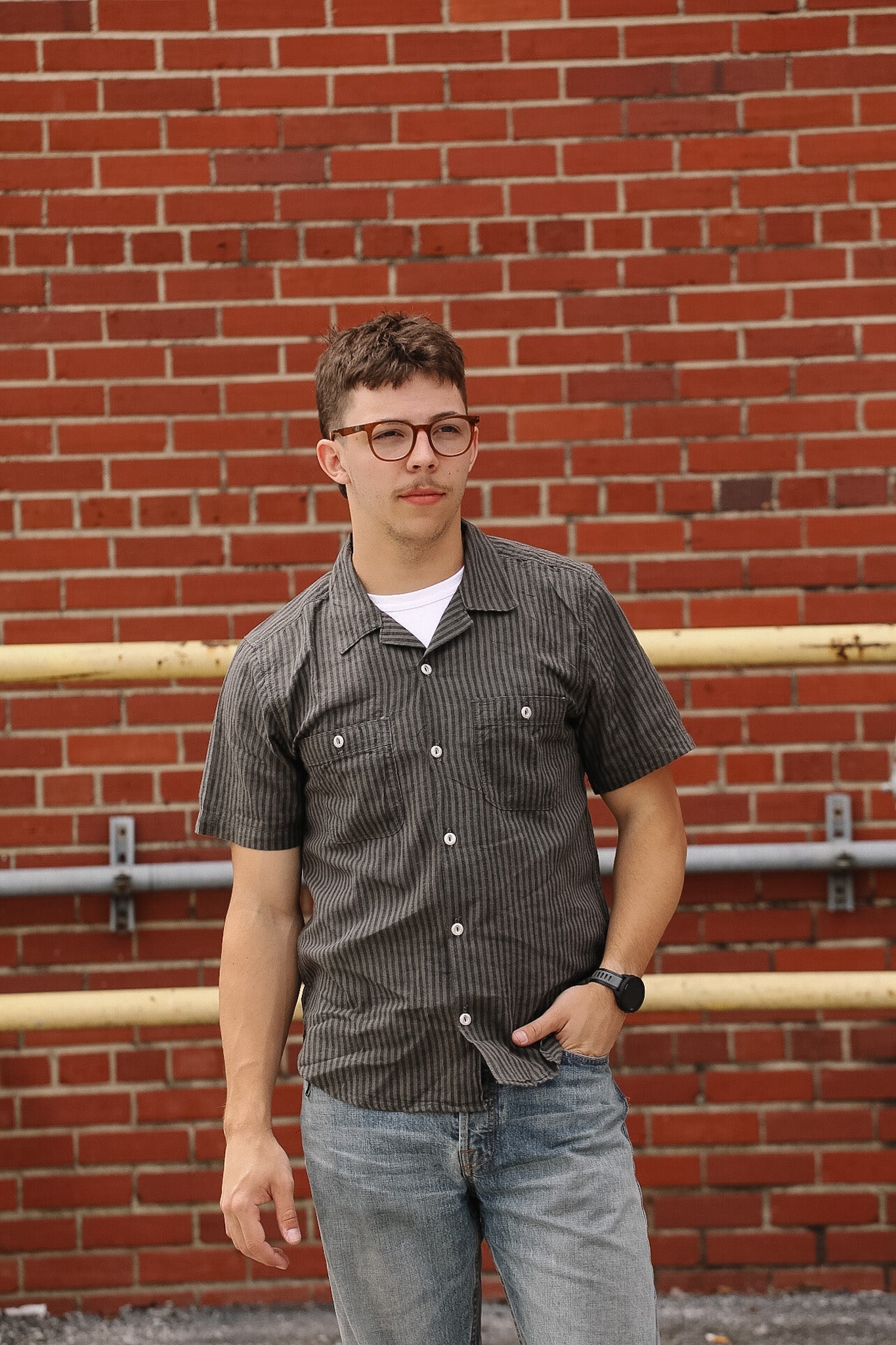 Man wearing a patterned shirt and jeans standing in front of a brick wall.