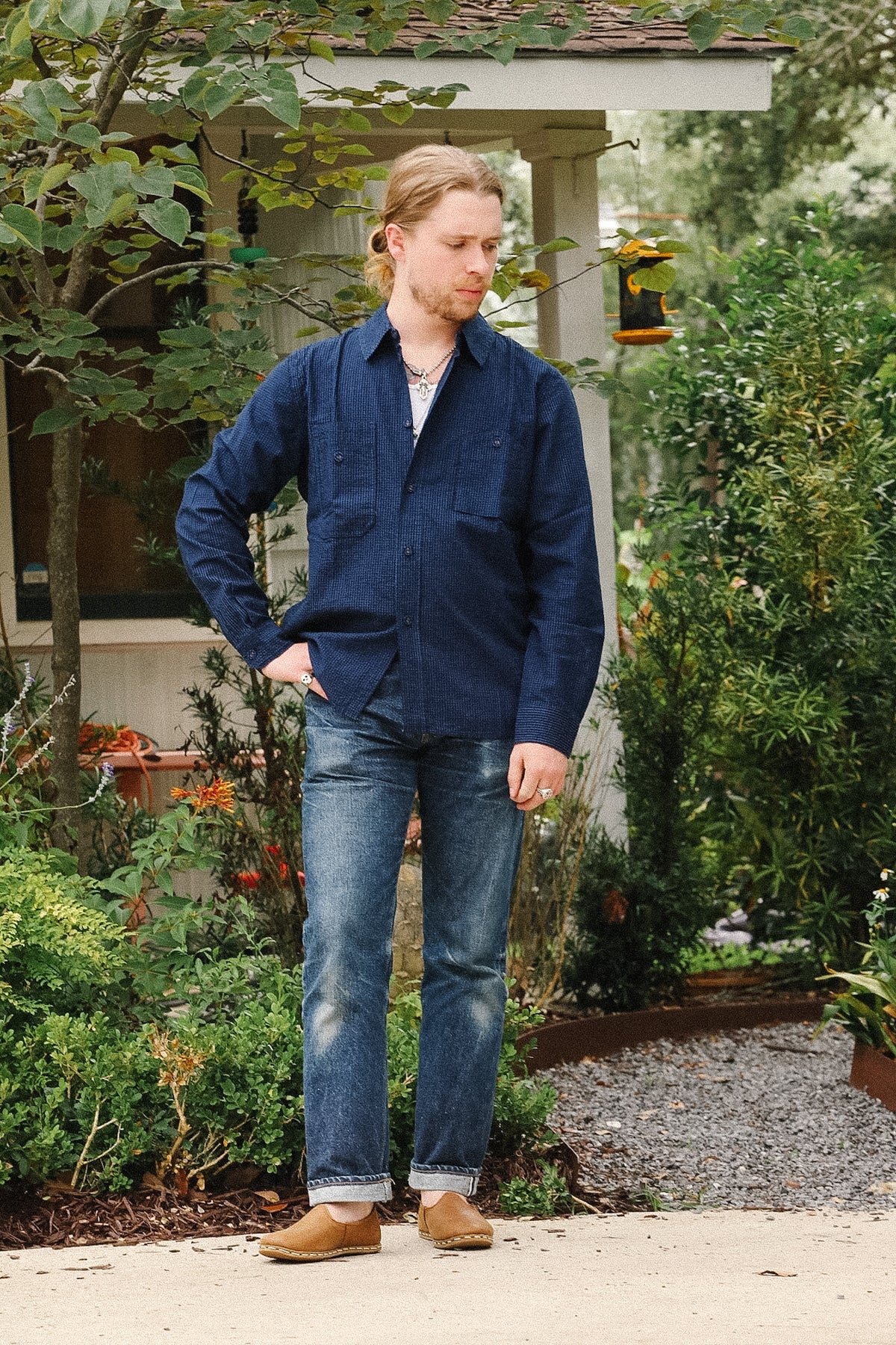 A man wearing a dark navy blue long-sleeve button-up shirt, blue jeans, and brown boots stands outdoors in front of a house with green garden plants.