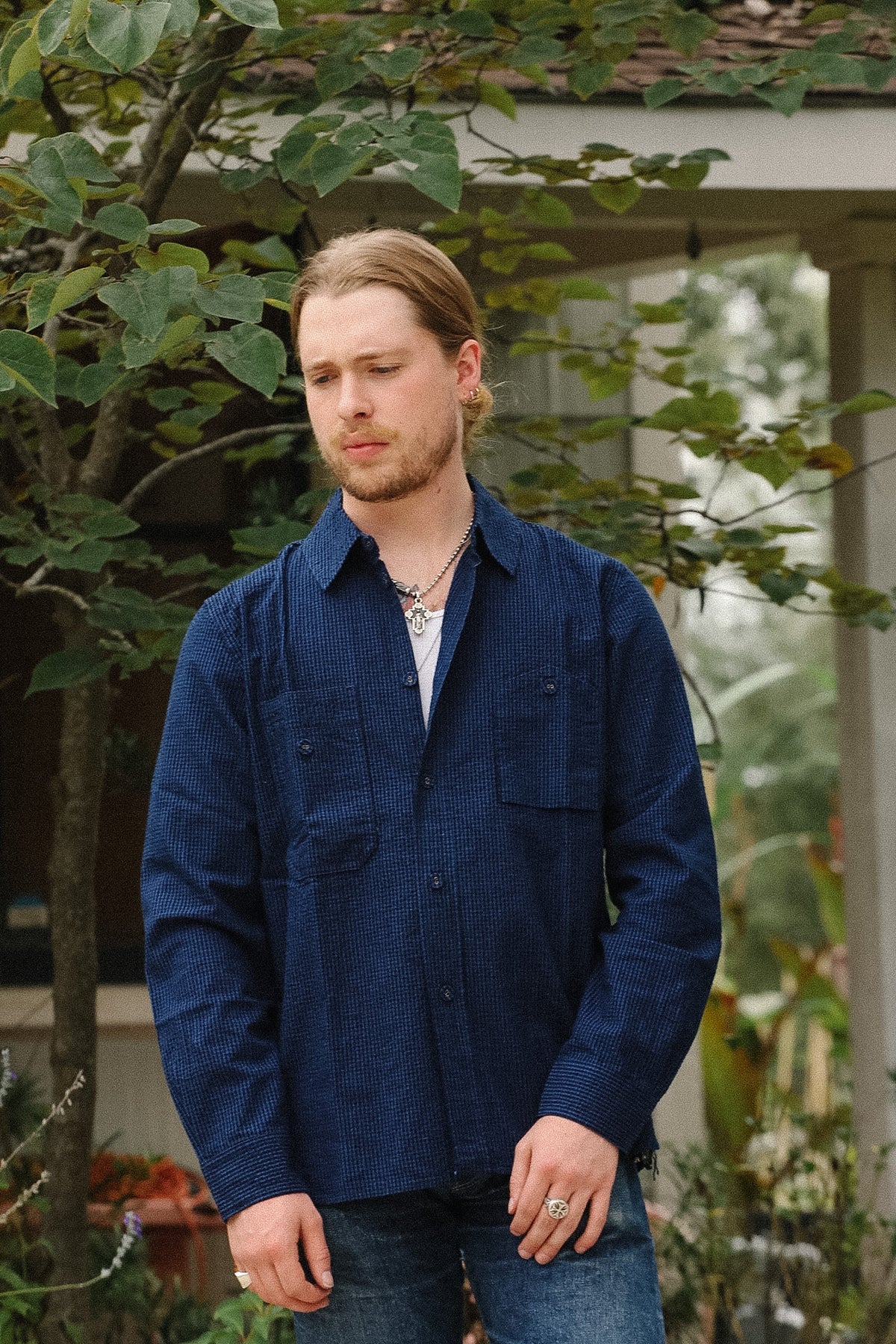 A man wearing a textured navy blue long-sleeve button-up shirt over a white undershirt, standing outdoors in front of a house with green foliage.
