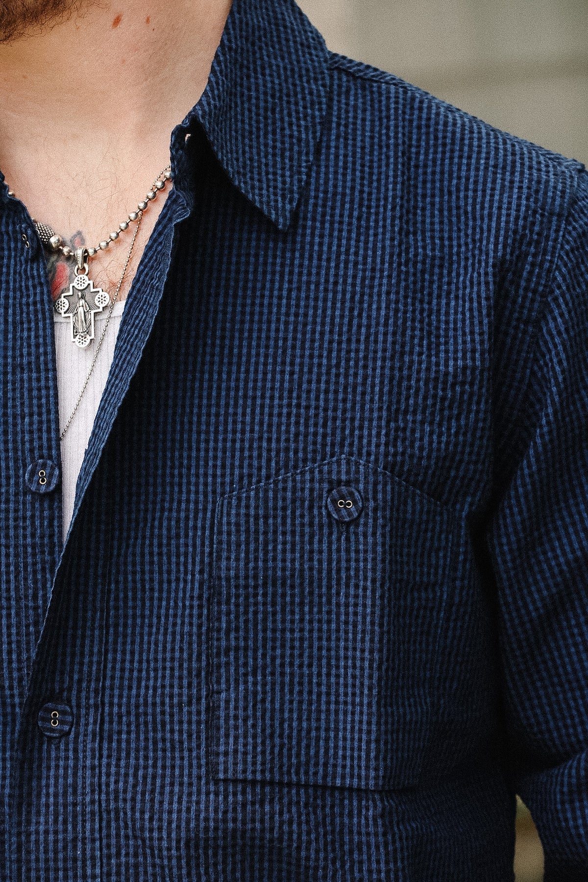 Close-up of a person wearing a dark blue and black micro-check patterned button-down shirt, layered over a white undershirt and accessorized with silver necklaces.