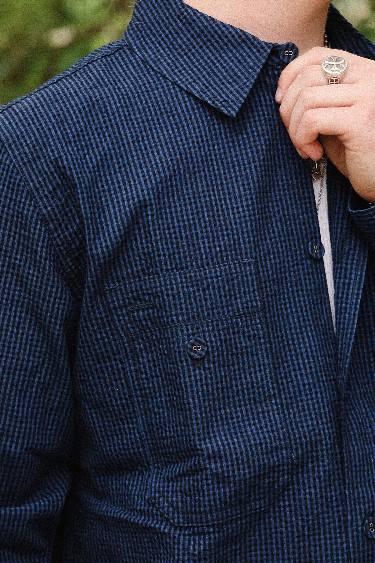 Close-up of a person wearing a dark navy blue and black micro-check patterned overshirt or jacket, with a hand adjusting the collar and a silver ring visible.