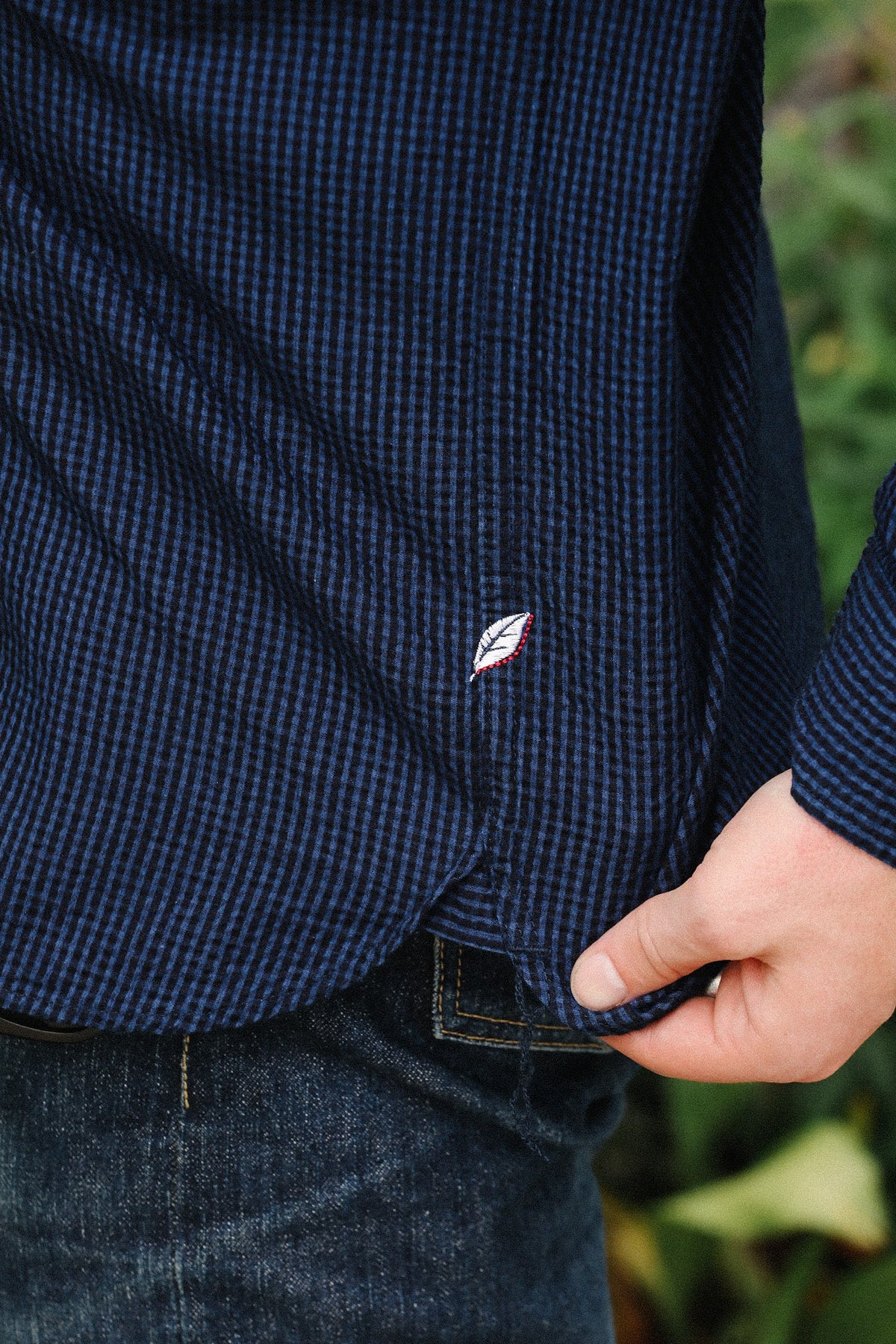 Close-up of a person wearing a dark navy blue and black micro-checkered long-sleeve button-down shirt, with a hand pulling the hem down slightly to reveal dark blue jeans underneath.