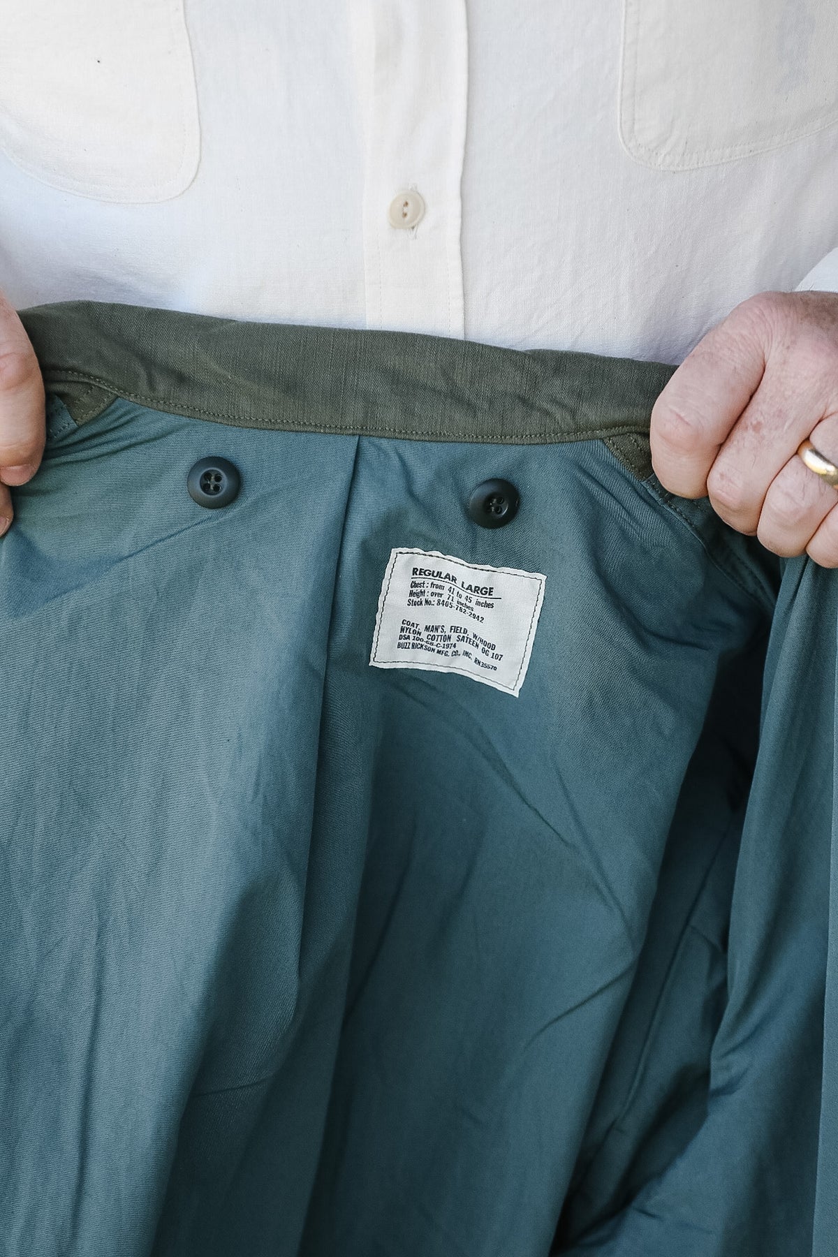 Green pants with a label being held open by hands, wearing a white shirt.