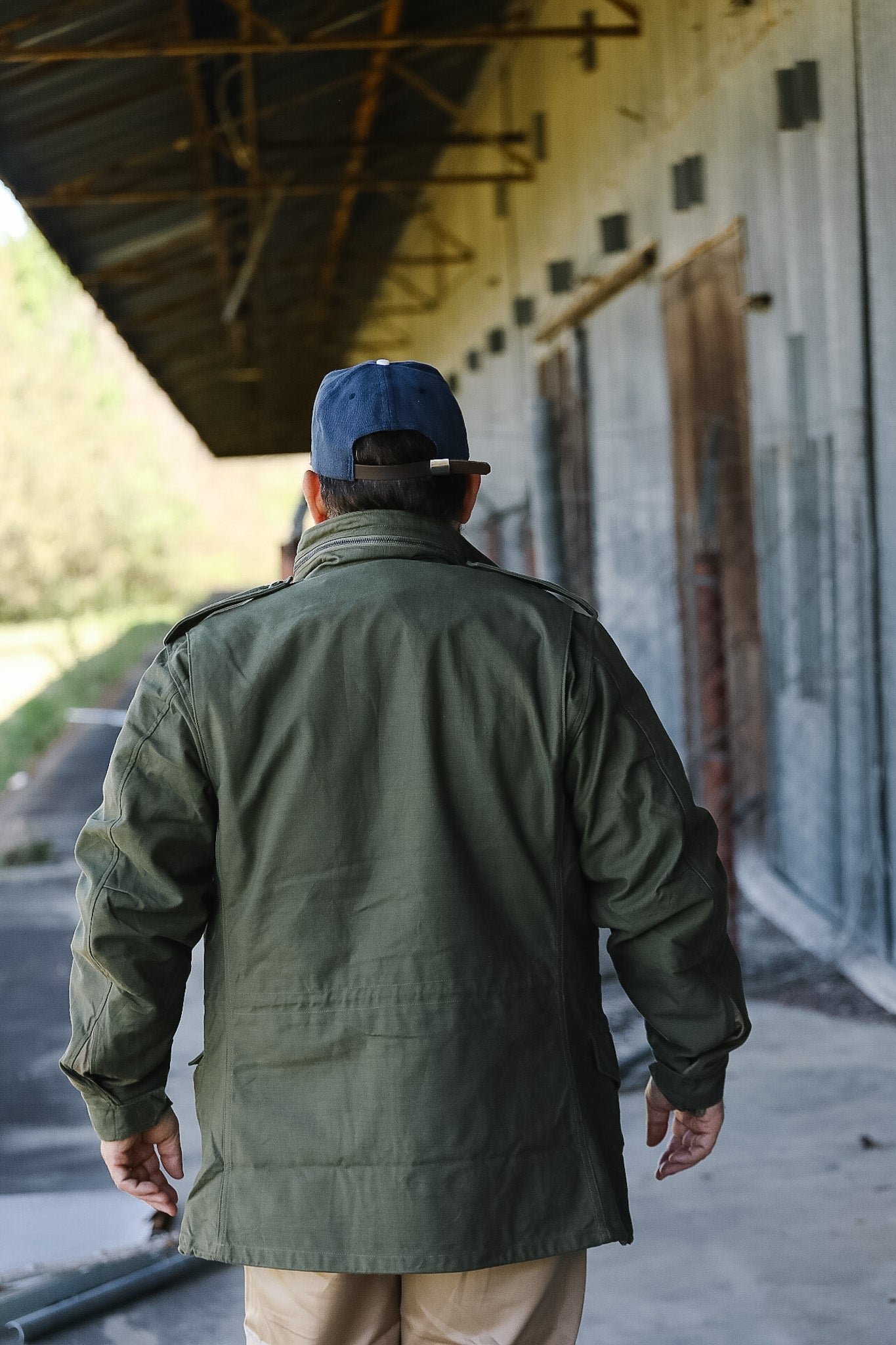 Person wearing a green jacket and blue cap walking away from the camera in an industrial setting.