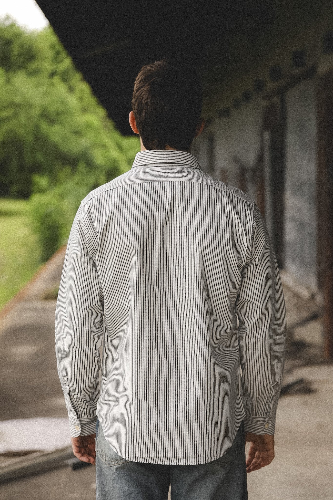 Man wearing a light gray striped shirt standing outdoors with greenery in the background