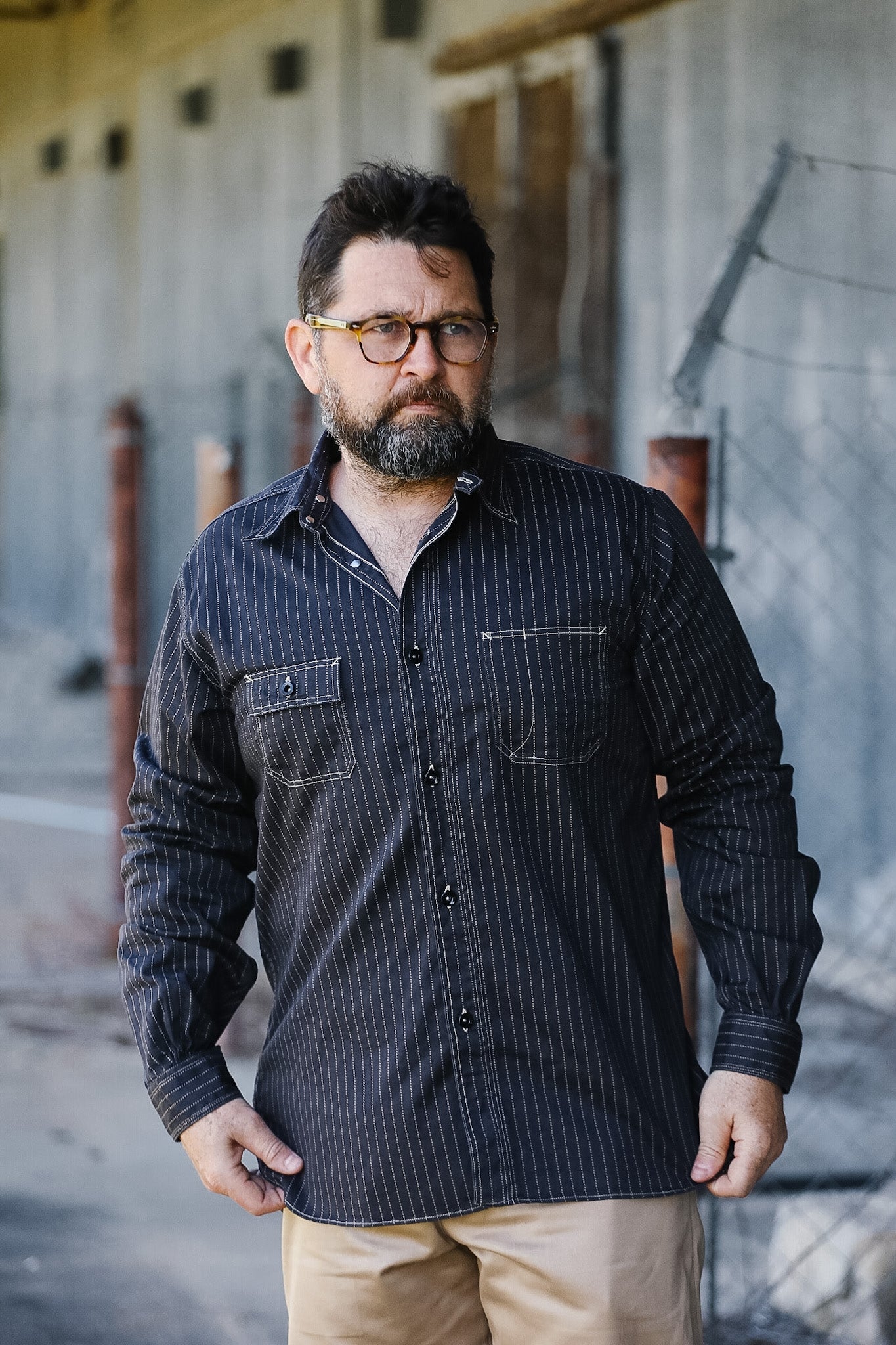 Man wearing a dark striped shirt and glasses standing in front of a concrete wall.