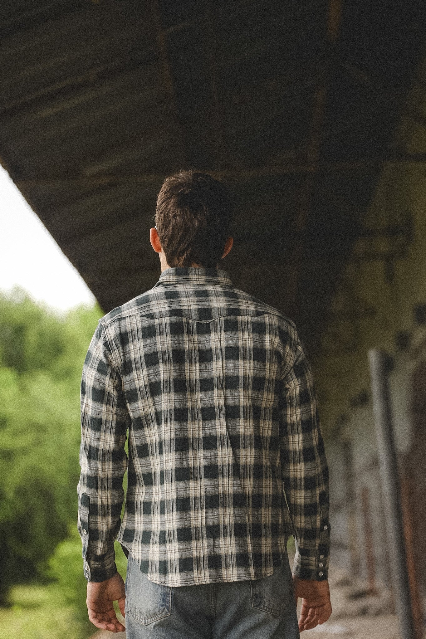 Person wearing a plaid shirt standing under a bridge with greenery in the background
