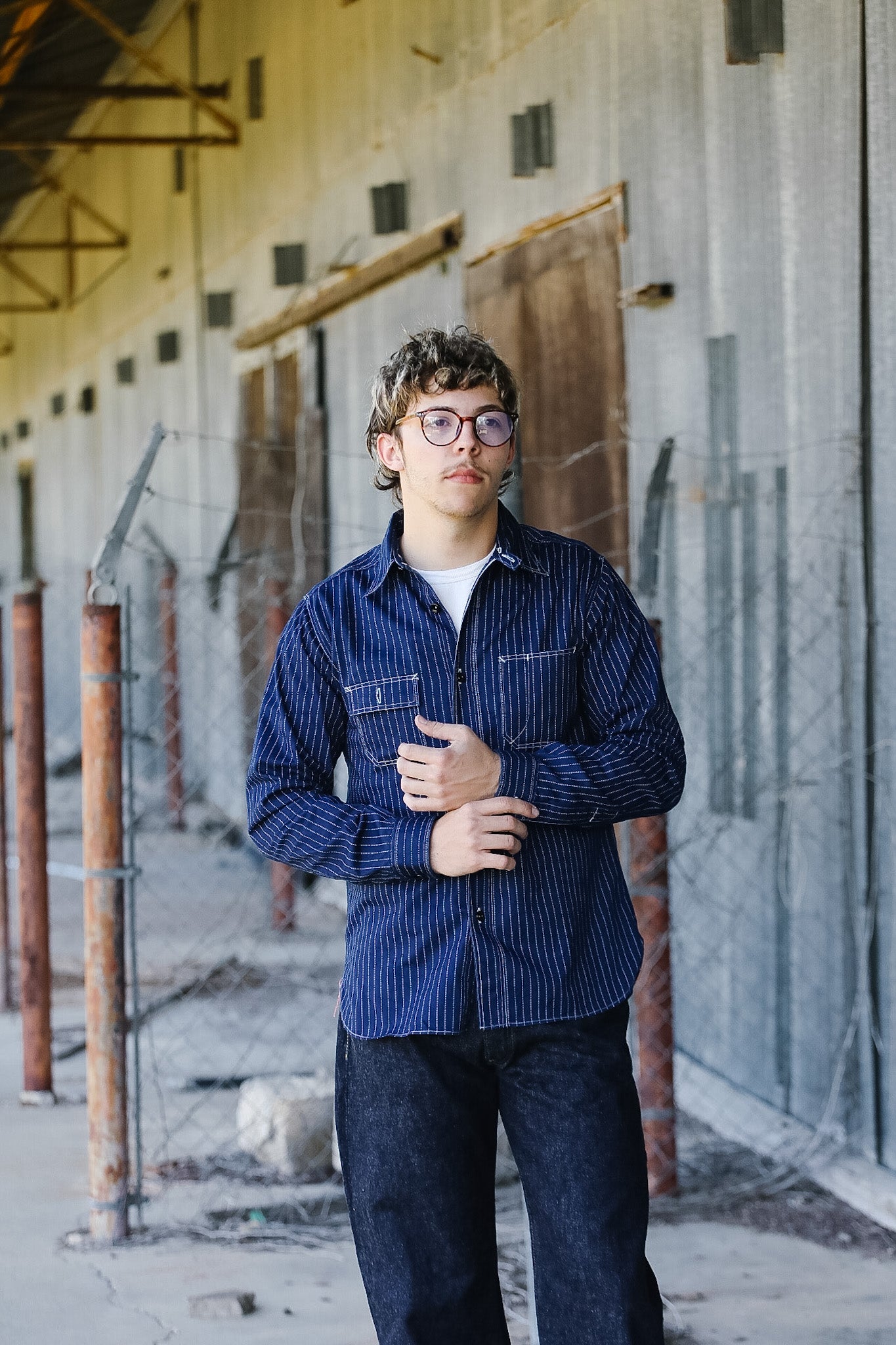 Person wearing a blue striped shirt and glasses standing in an industrial setting.