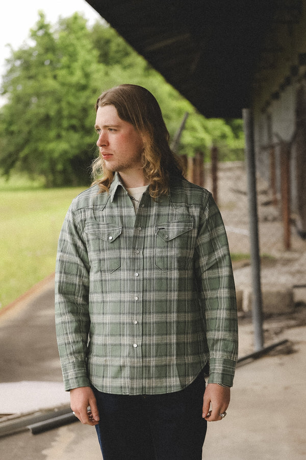 Person wearing a green plaid shirt standing outdoors with trees and a building in the background.
