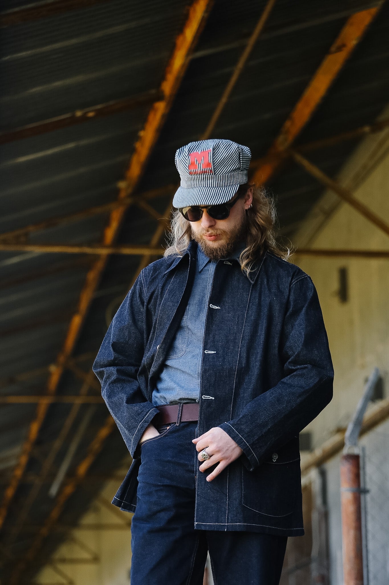 Man wearing a denim jacket and cap with a logo, standing in an industrial setting.
