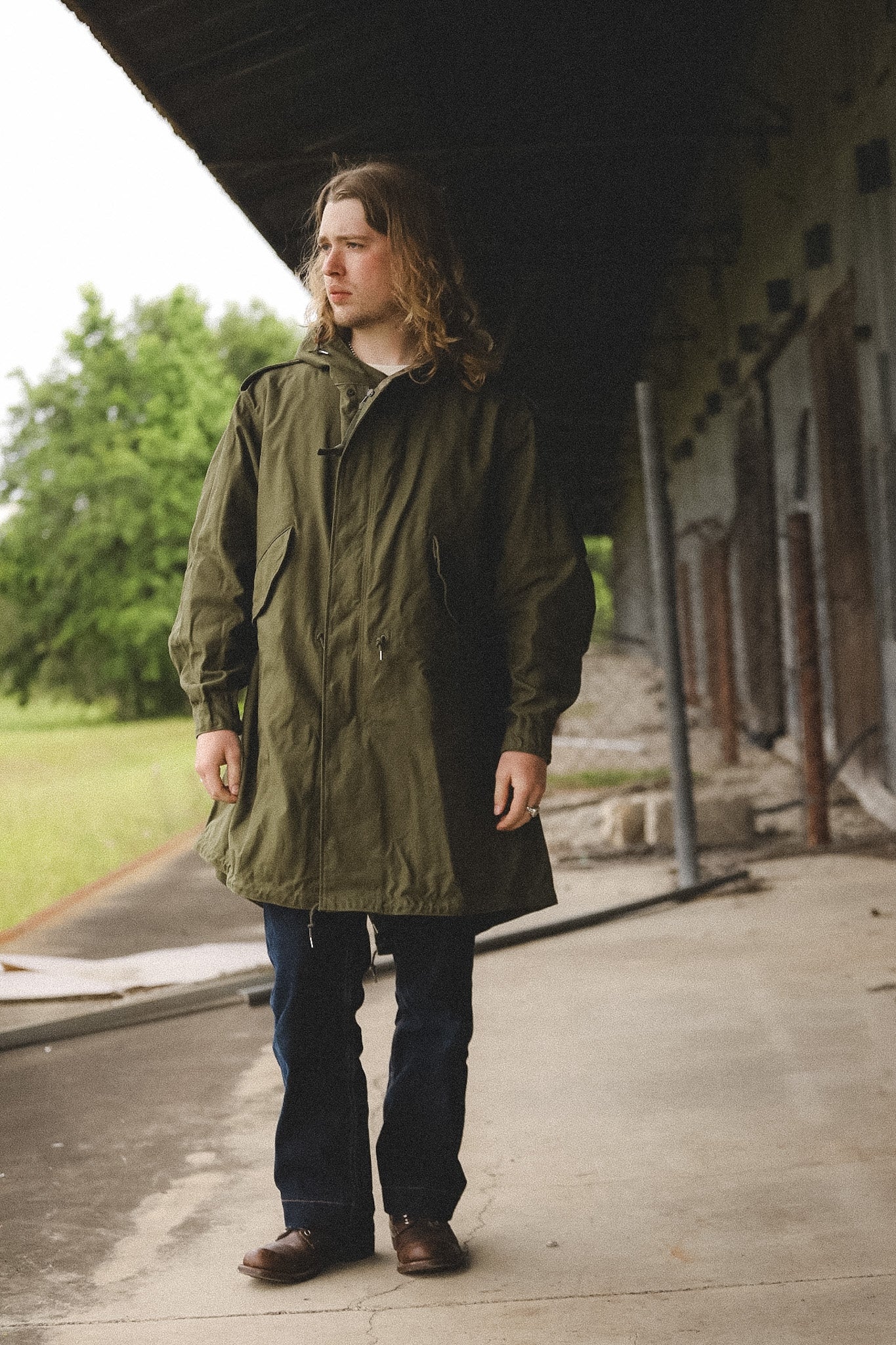 Man wearing a green coat standing on a wooden platform with a building and trees in the background