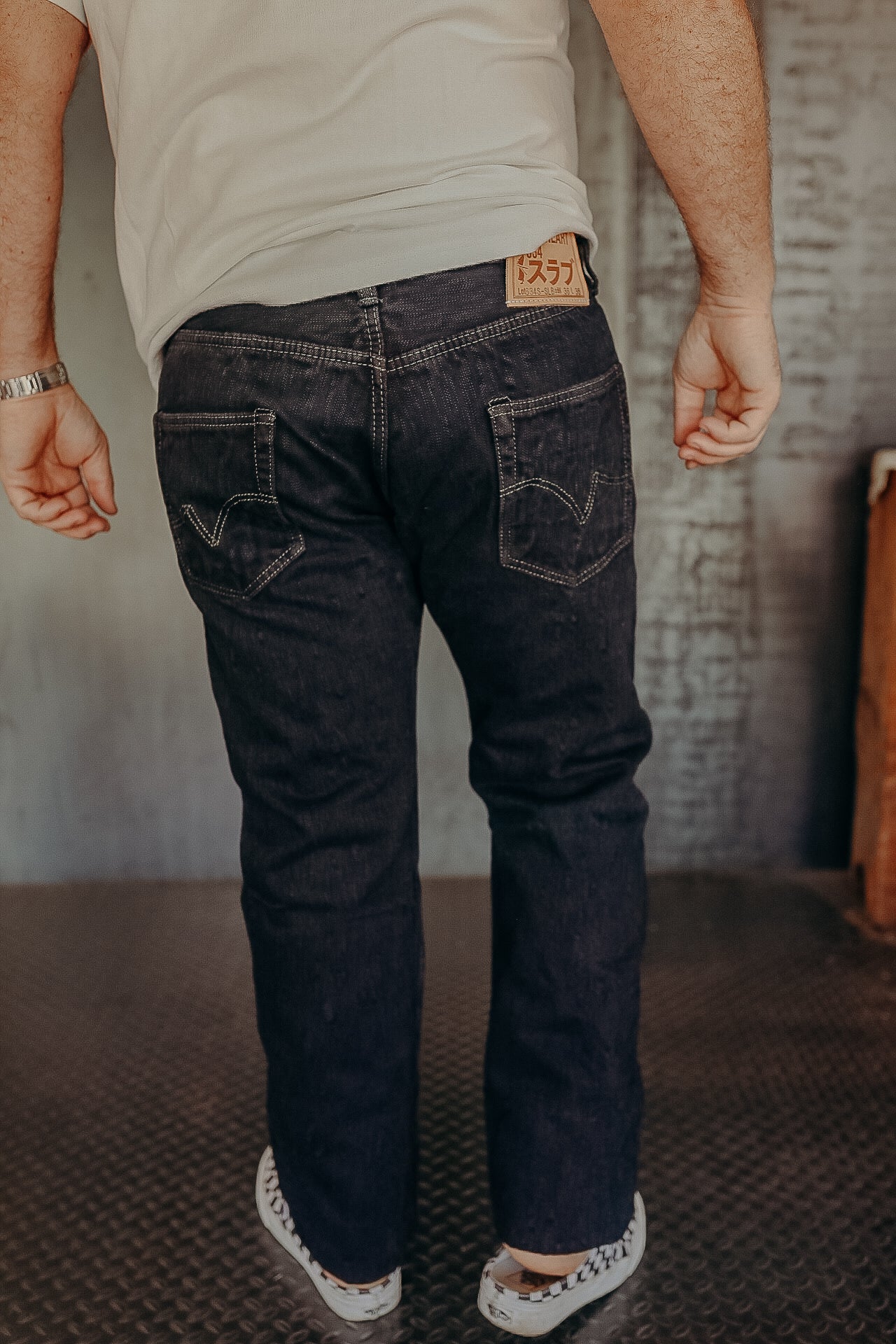 Man wearing dark denim jeans with double-stitched back pockets and a white shirt, standing indoors