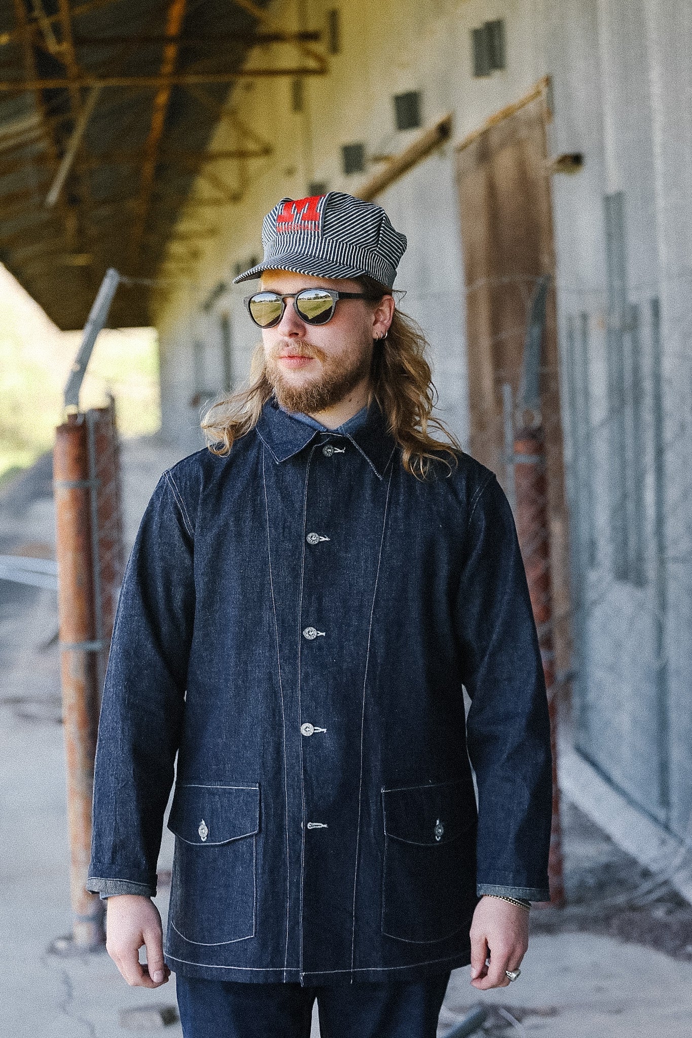 Man wearing a dark denim coat and sunglasses standing in an industrial setting.
