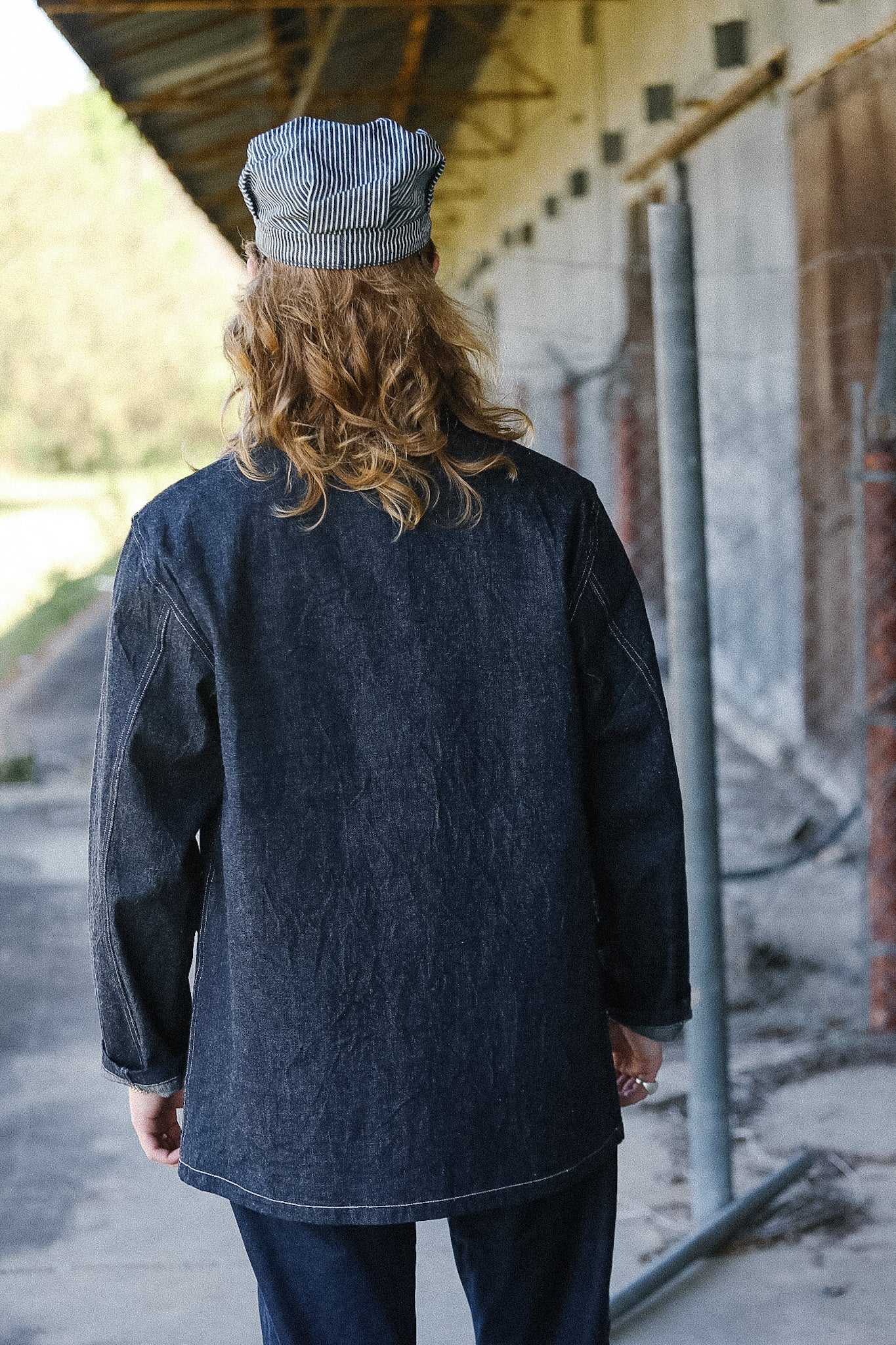 Person wearing a dark denim jacket and cap standing in an outdoor setting with a building in the background.
