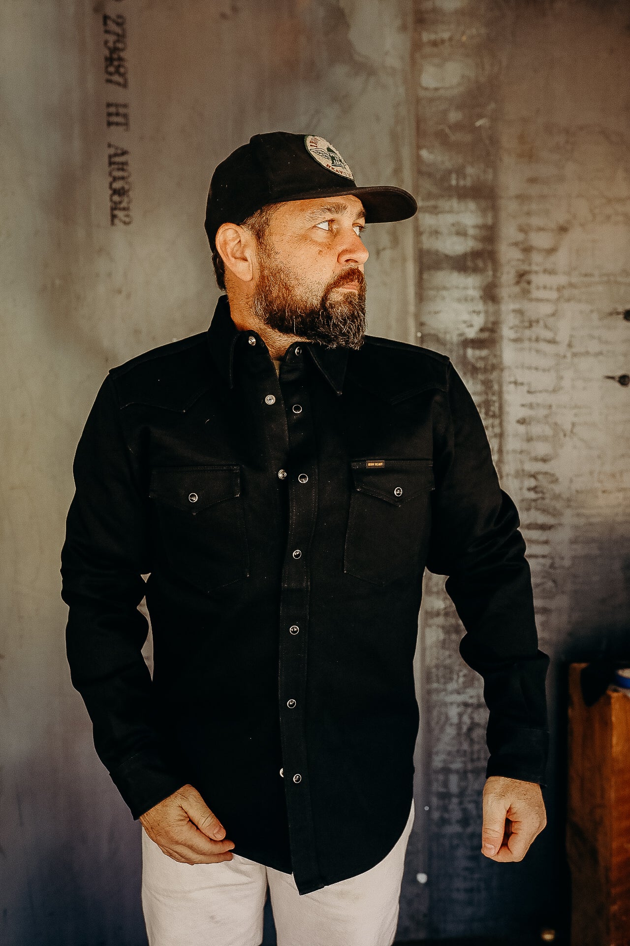 Man with beard wearing black work shirt and cap standing against textured concrete wall