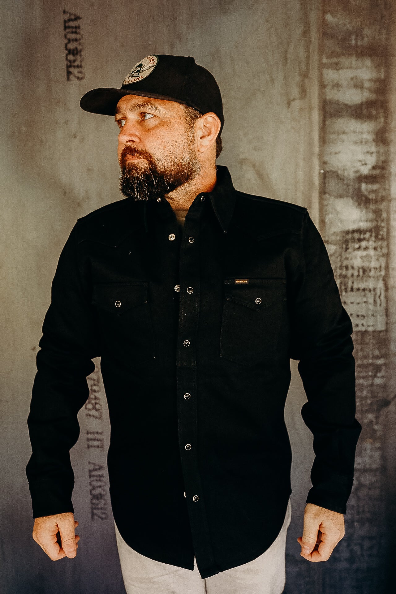 Man wearing black Iron Heart jacket and black cap with patch standing against textured wall