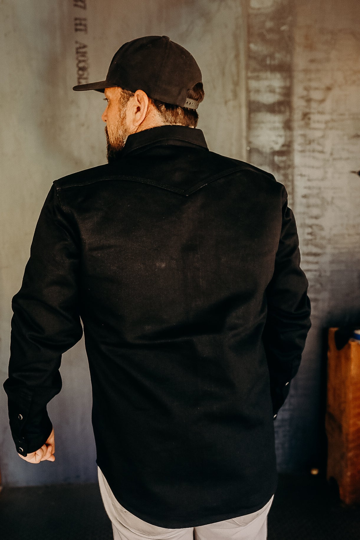 Man wearing black work shirt and black cap standing indoors against industrial wall