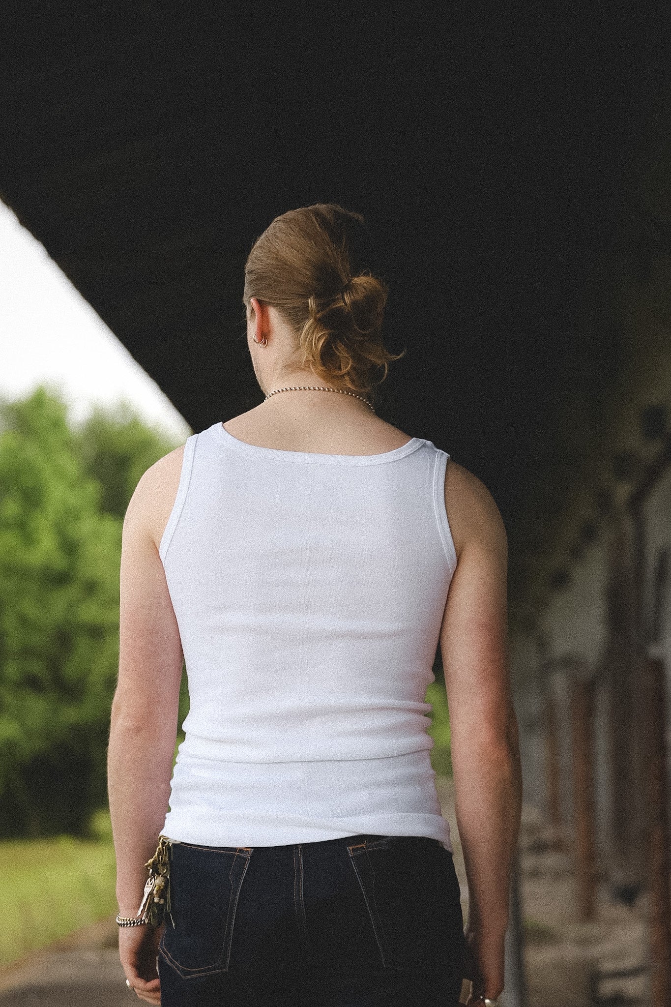 Person wearing a white tank top and dark pants standing outdoors with trees and a building in the background.