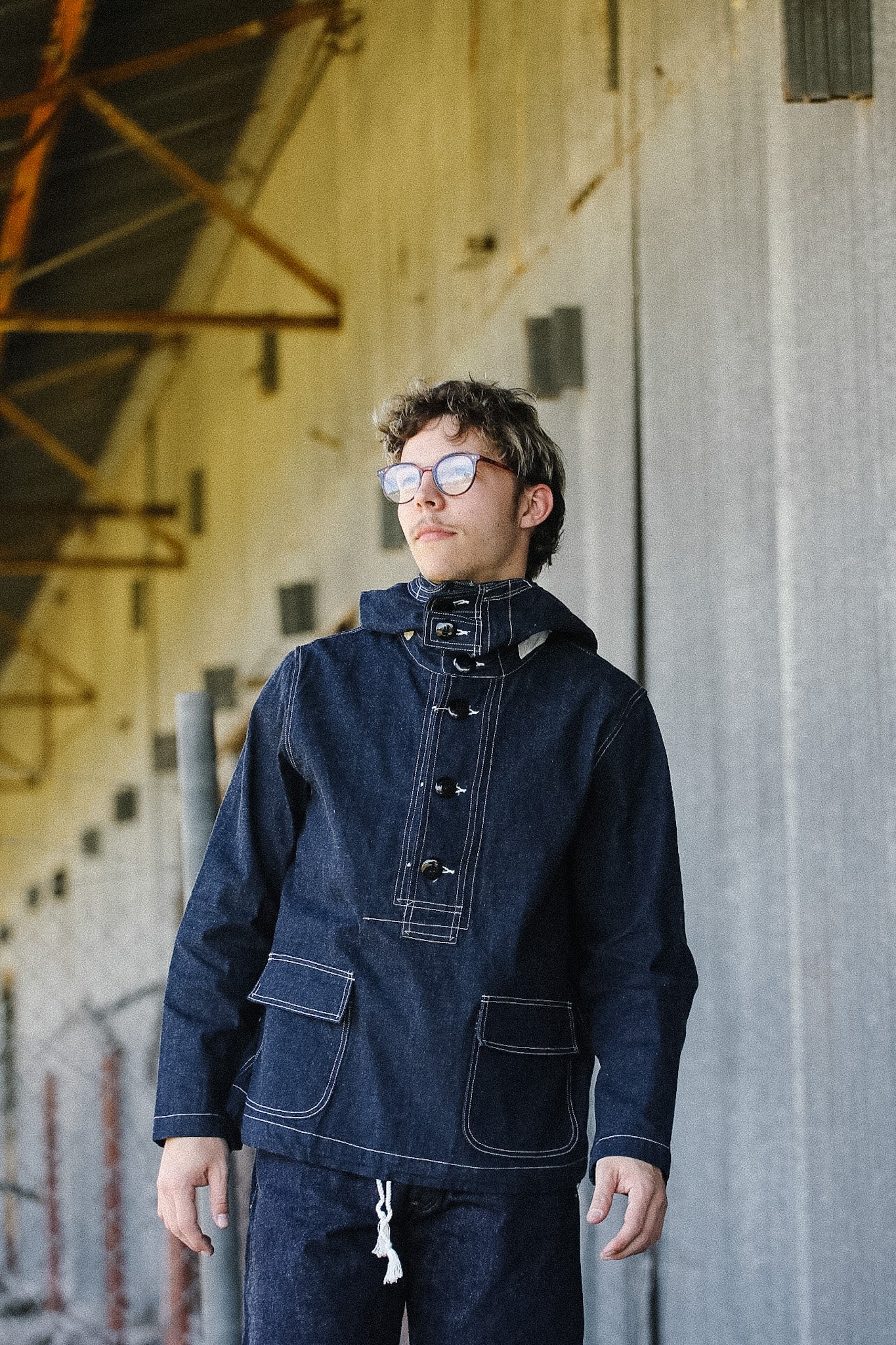 Person wearing a dark denim jacket and pants standing against a textured wall.