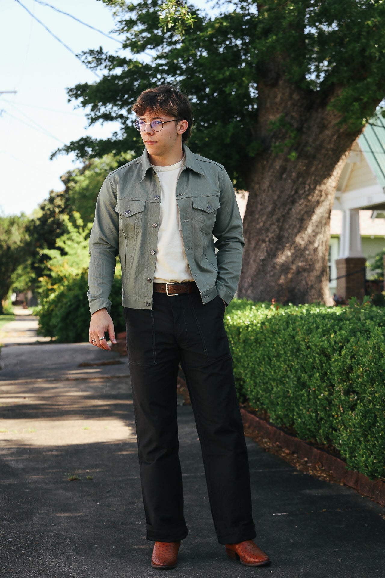 Man wearing green jacket, white shirt, black pants, and brown boots standing on sidewalk with large tree and greenery