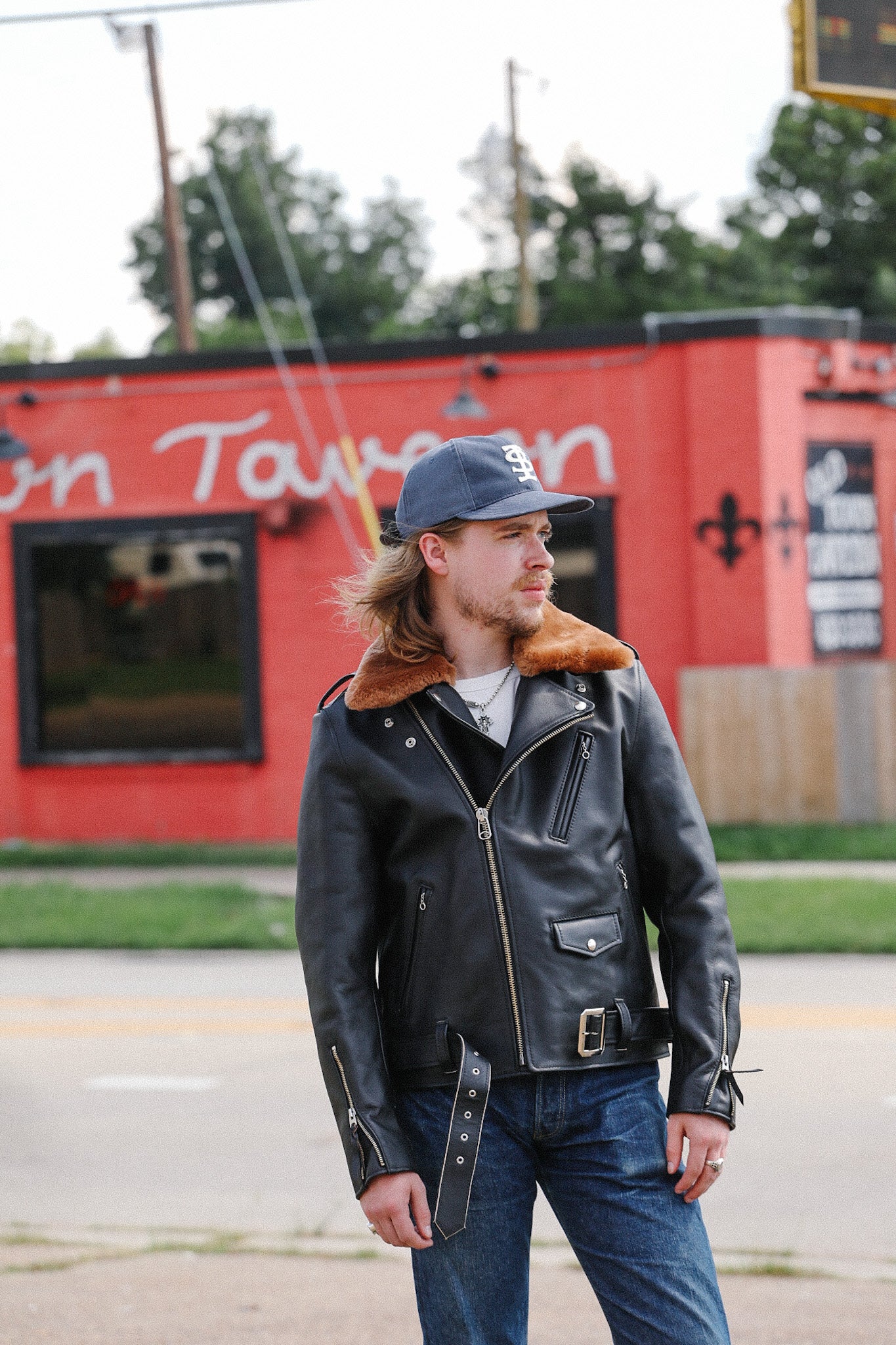 Man wearing a black leather jacket with fur collar and blue baseball cap standing outdoors