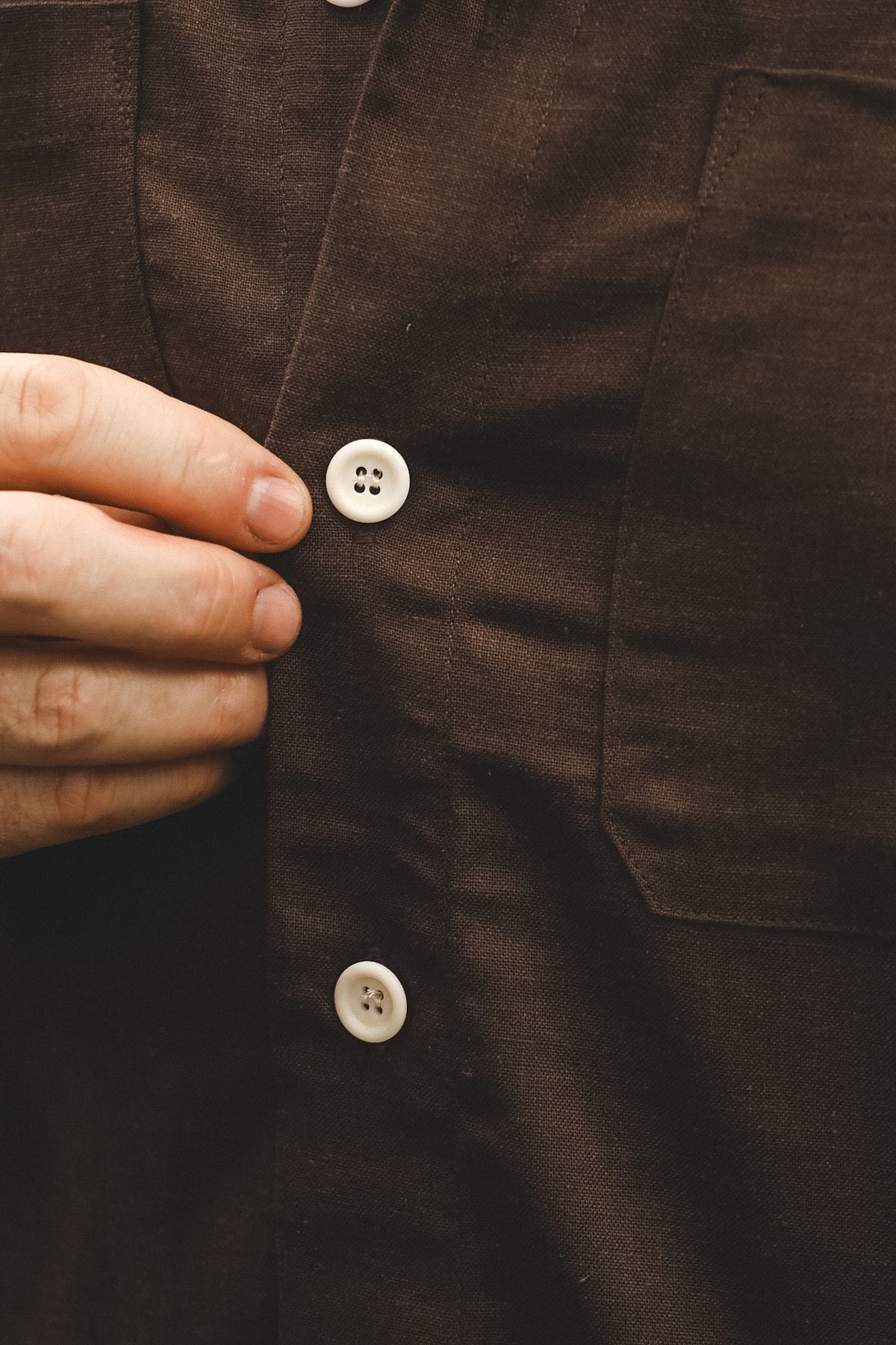 Close-up of a dark brown linen button-up shirt being fastened by a hand, showing white buttons and the textured fabric.