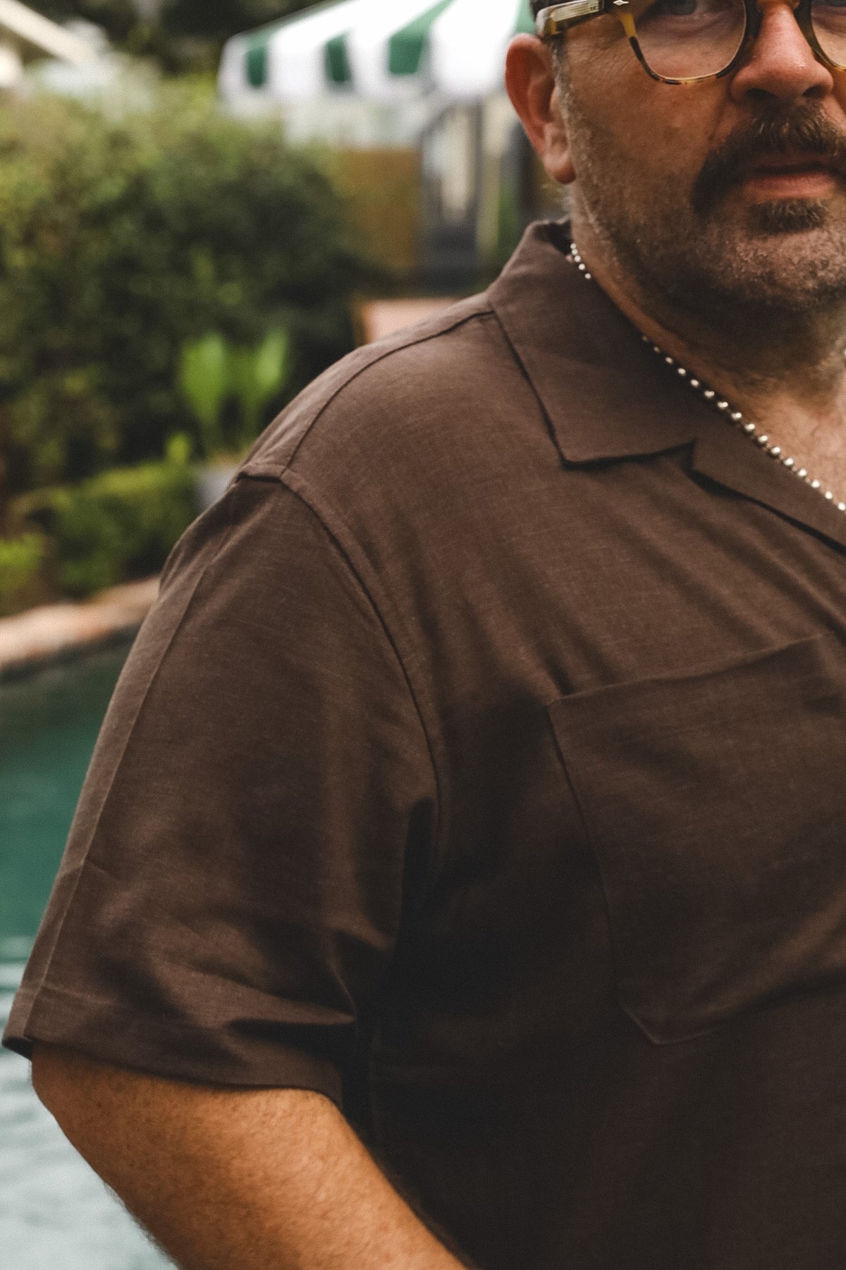 A close-up shot of a man wearing a dark brown short-sleeve button-down shirt with a chest pocket, standing outdoors near a pool.
