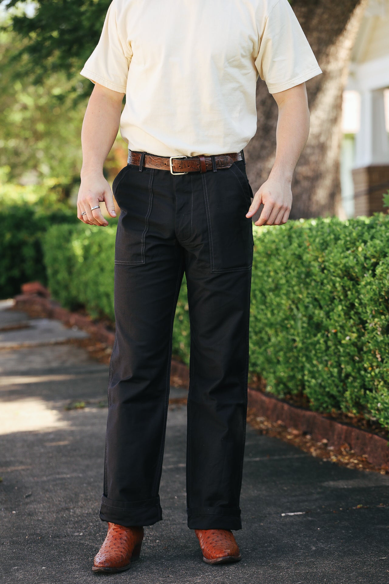 Man wearing black work pants, brown leather belt, cream t-shirt, and brown leather boots standing outdoors