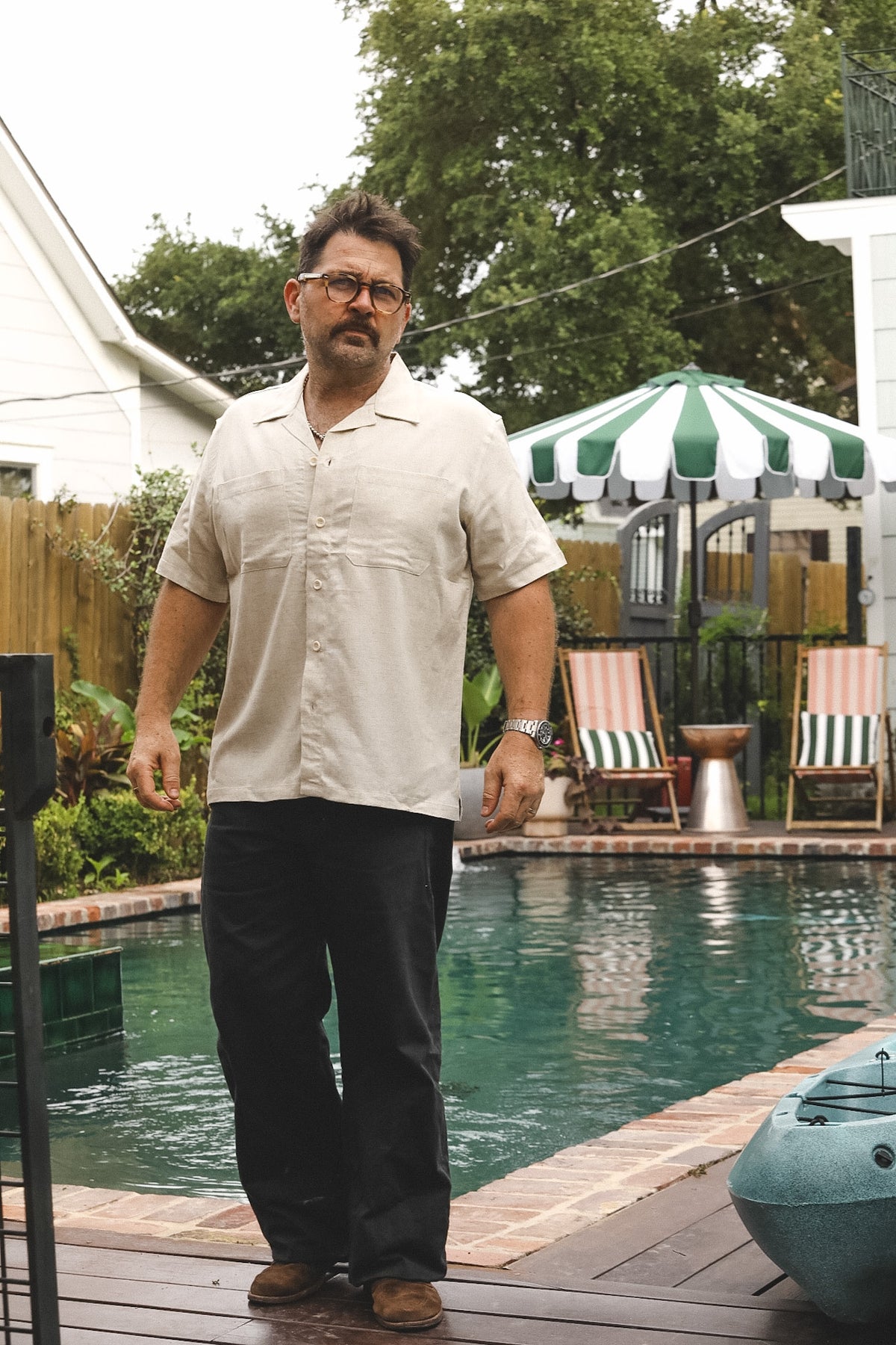 A man wearing a beige short-sleeve button-up shirt and dark pants stands on a wooden deck next to a swimming pool with patio chairs and a green-and-white striped umbrella in the background.
