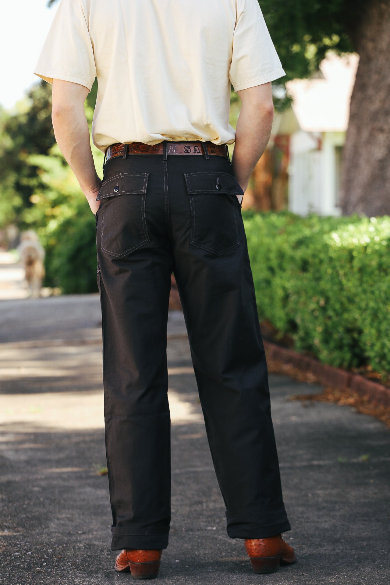 Rear view of person wearing black work pants with buttoned back pockets, tan shirt, brown belt, and boots on sidewalk