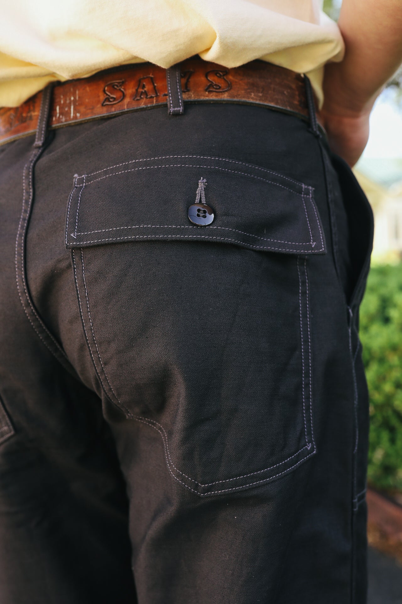 Close-up of black durable work pants pocket with button flap, worn with brown leather belt outdoors