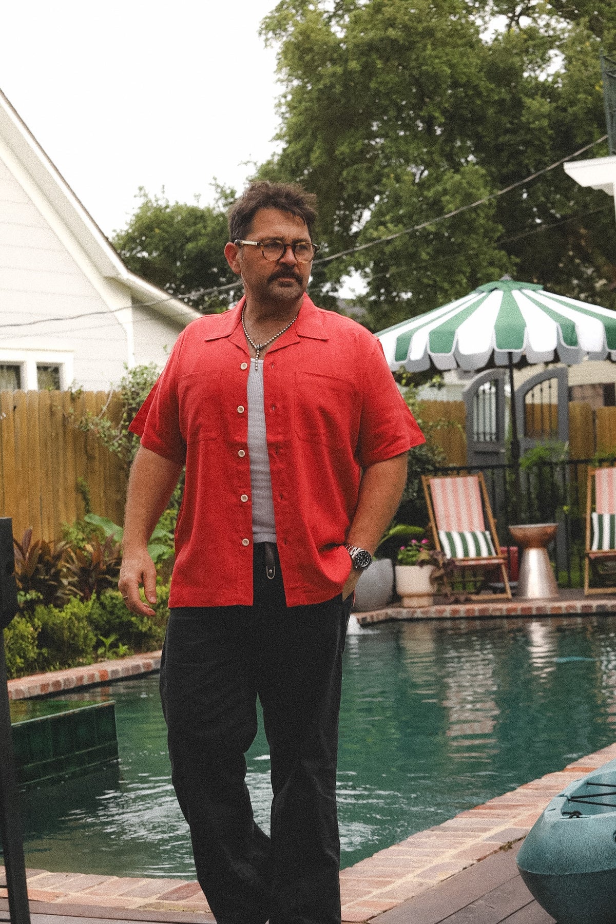 A man wearing a red short-sleeve button-up shirt, black pants, and glasses stands next to a swimming pool in a backyard with a green and white striped patio umbrella and lounge chairs.