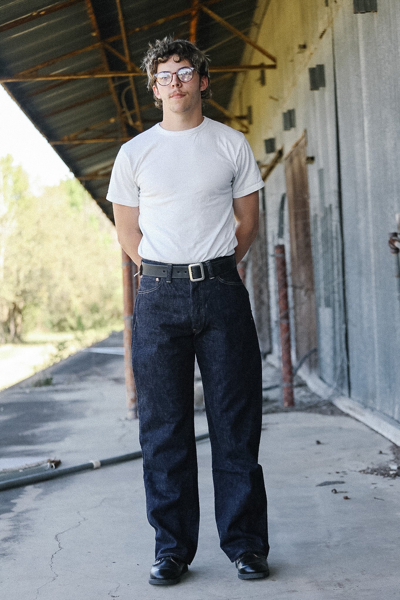 Man wearing a white t-shirt and dark jeans standing in an outdoor setting with a building in the background.