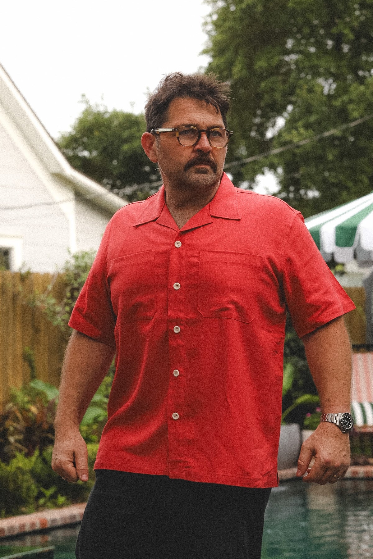 A man wearing a bright red short-sleeve camp collar button-up shirt and black pants, standing outdoors near a swimming pool with trees and a house in the background.