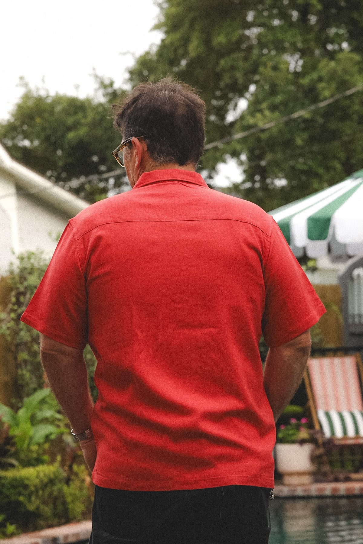 A man viewed from behind wearing a bright red short-sleeve button-up shirt and dark pants, standing outdoors near a pool with trees and patio furniture in the background.