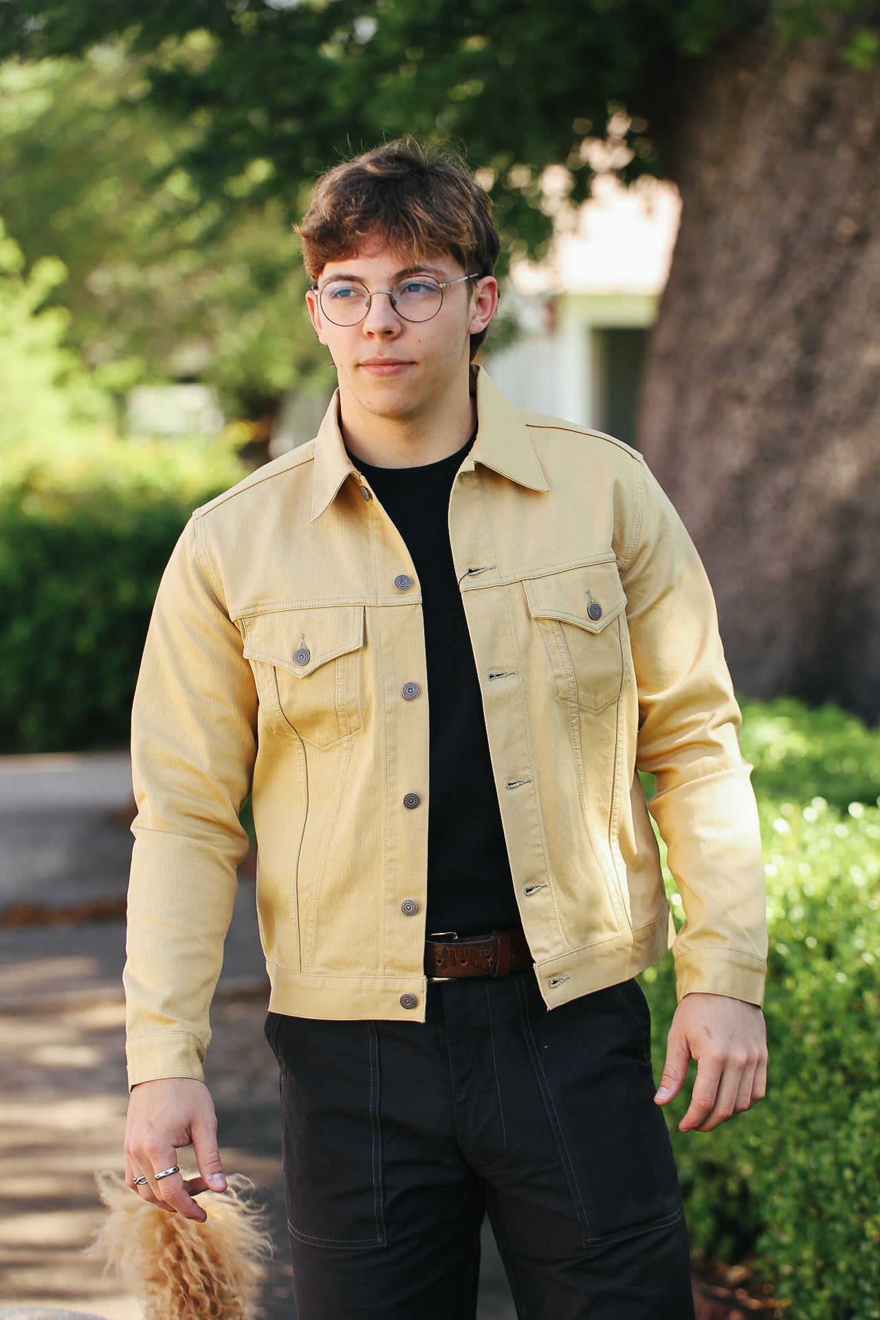 Young man wearing beige jacket and black pants standing outdoors near green bushes and tree