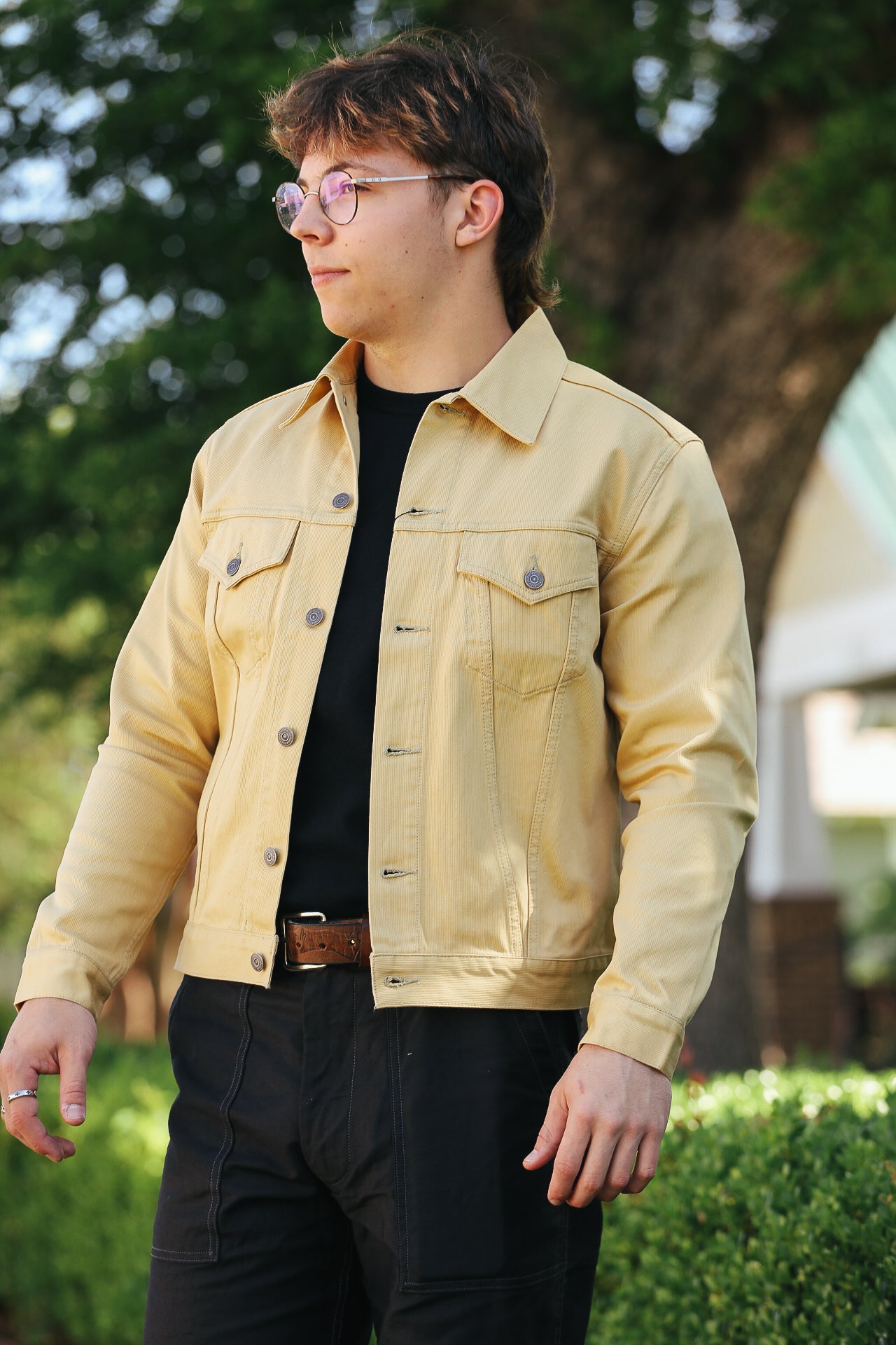 Man wearing a tan workwear jacket, black shirt, and black work pants standing outdoors near greenery
