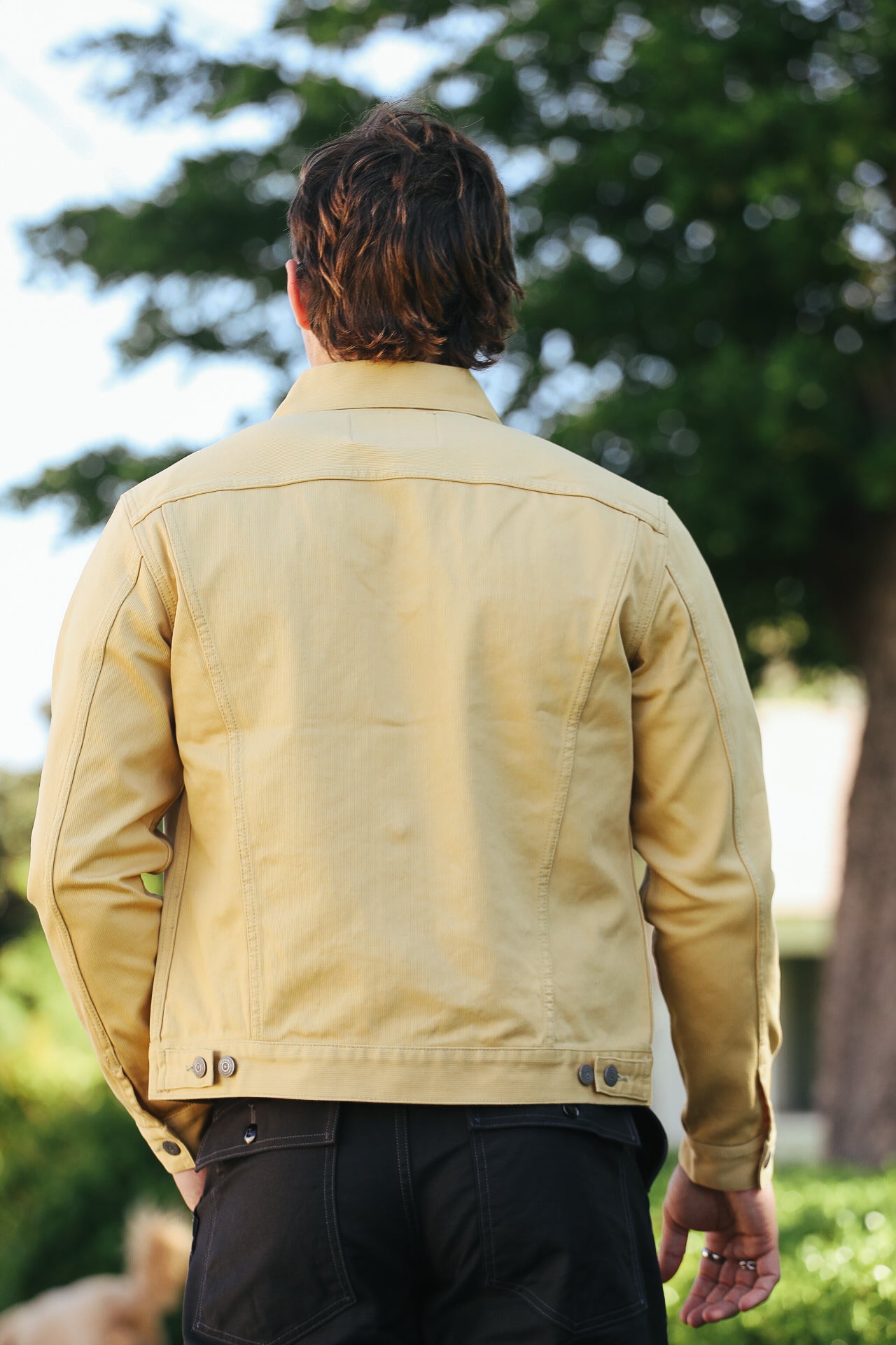 Man wearing yellow workwear jacket with button details standing outdoors with greenery background