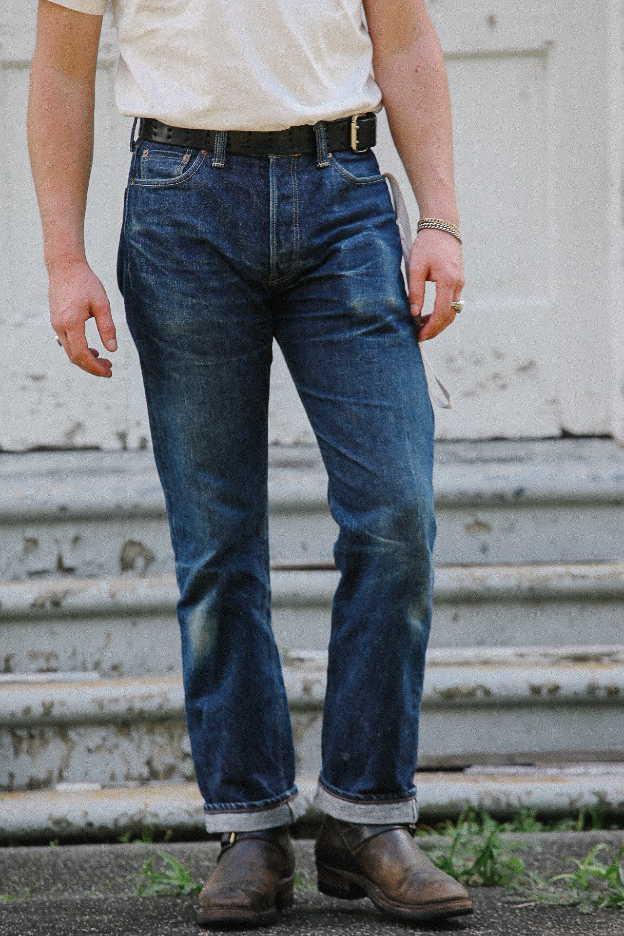 Man wearing dark blue denim jeans with rolled cuffs, brown leather boots, black belt, and white shirt standing by worn concrete steps