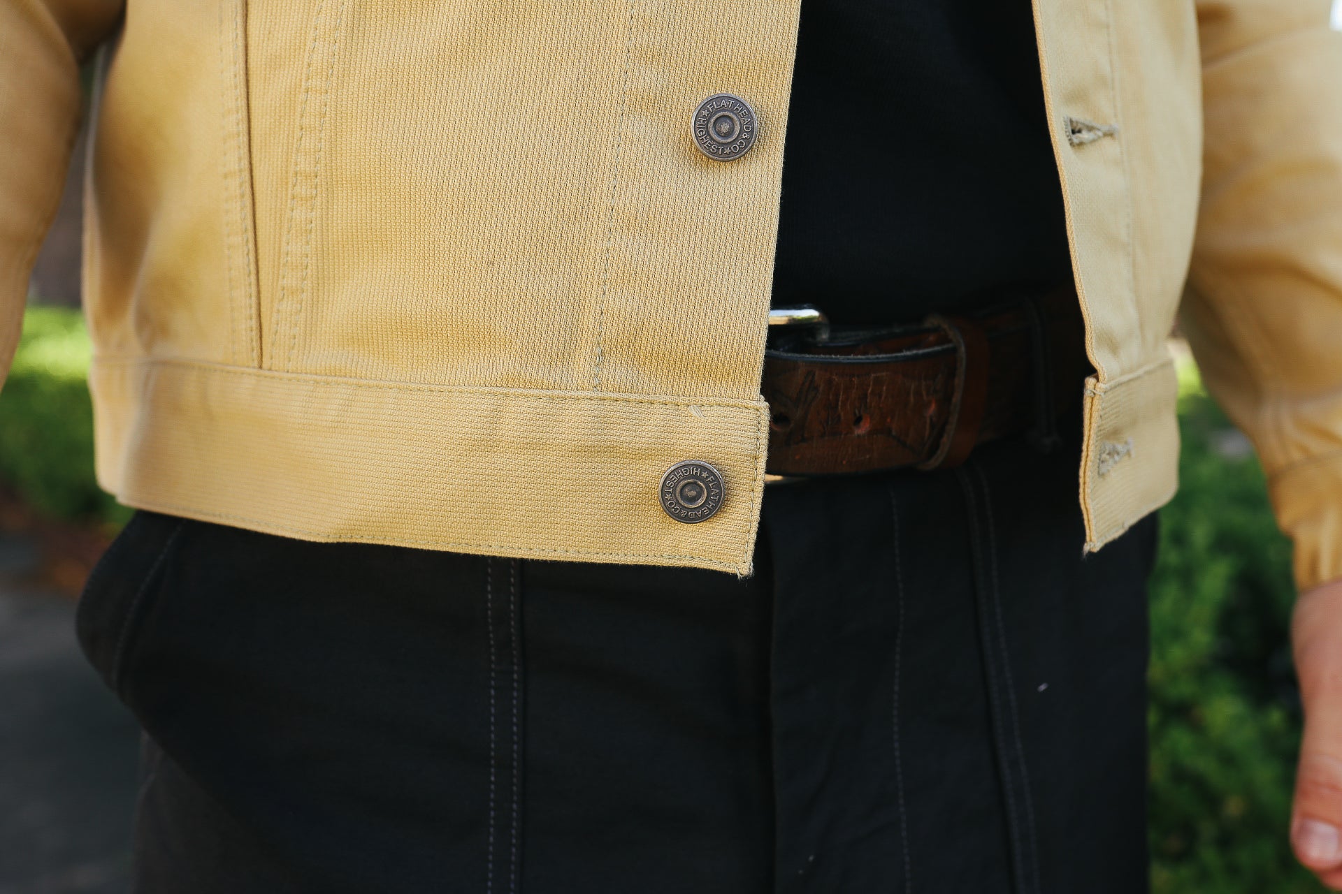 Close-up of person wearing a vintage yellow work jacket with metal buttons and black pants with a brown leather belt