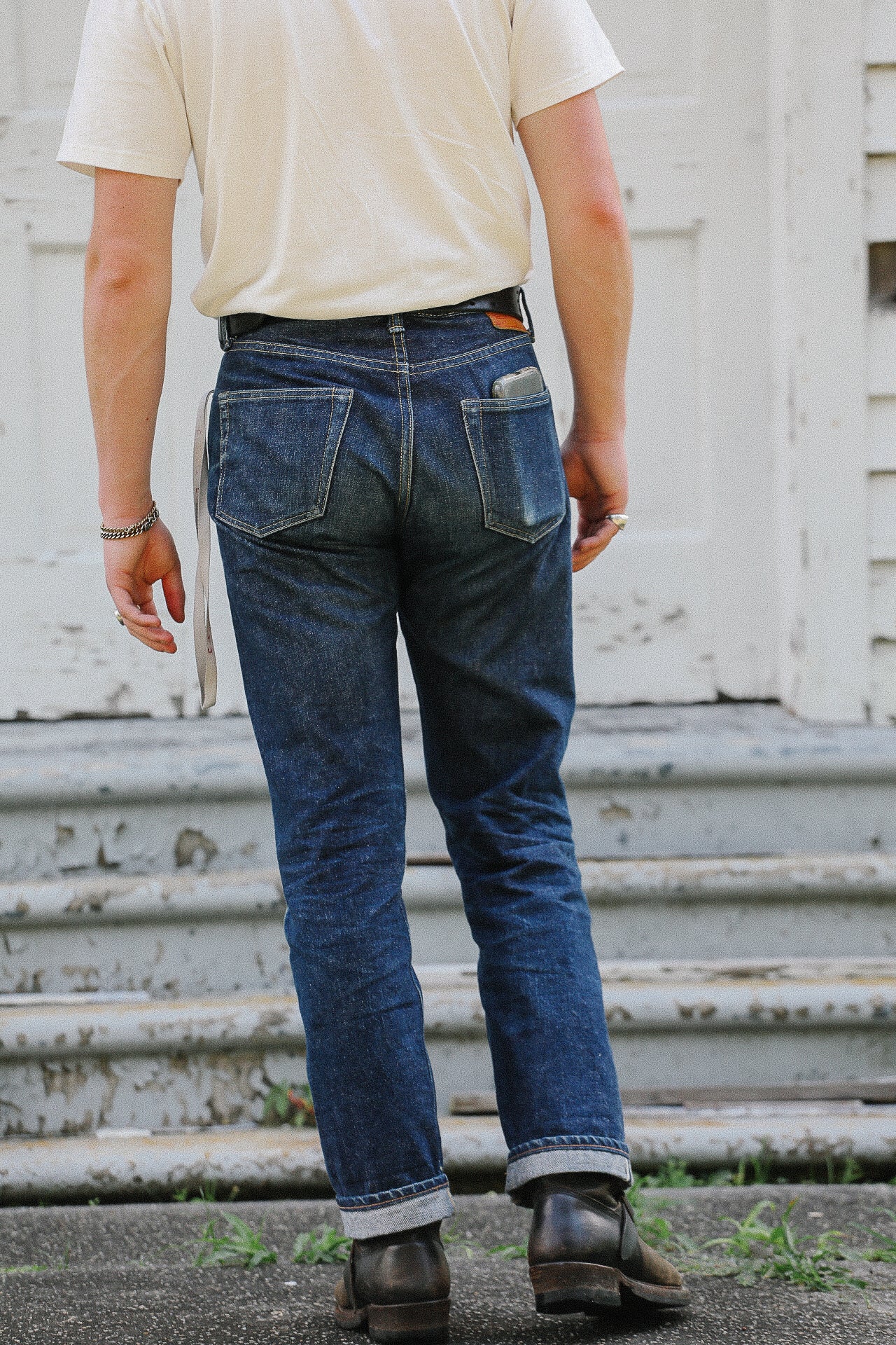 Back view of person wearing cuffed dark denim jeans, black leather boots, and a cream t-shirt standing on worn steps