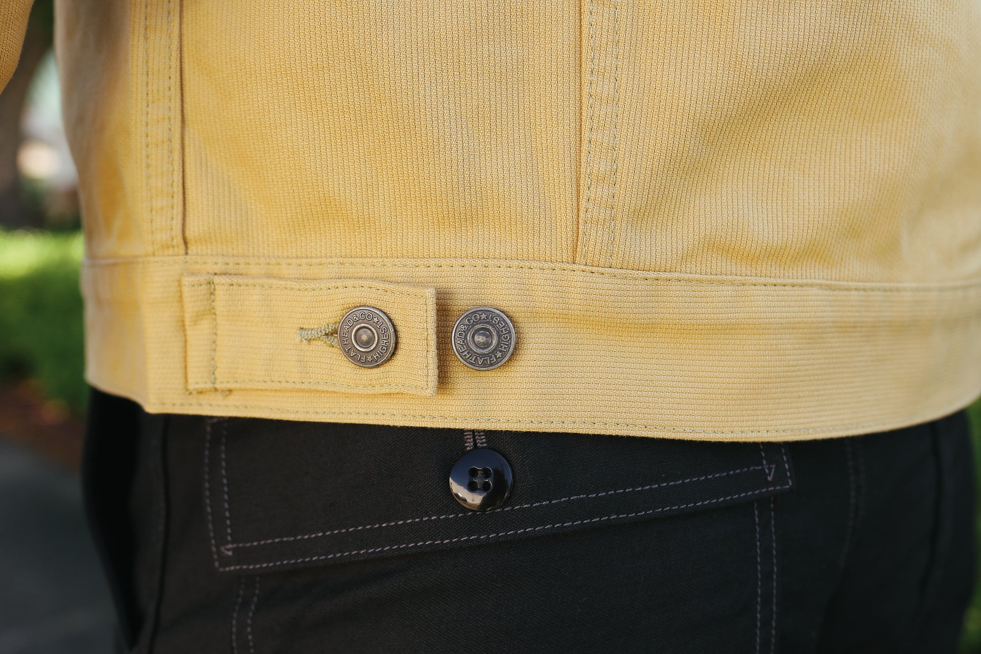 Close-up of yellow denim jacket hem with metal buttons over black pants with white stitching