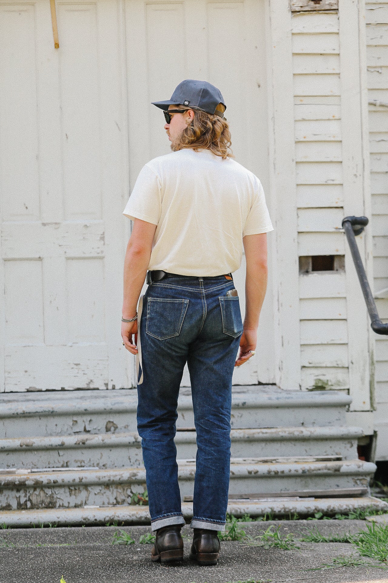 Man wearing cuffed dark denim jeans, white t-shirt, navy cap, and boots standing by weathered white wooden building