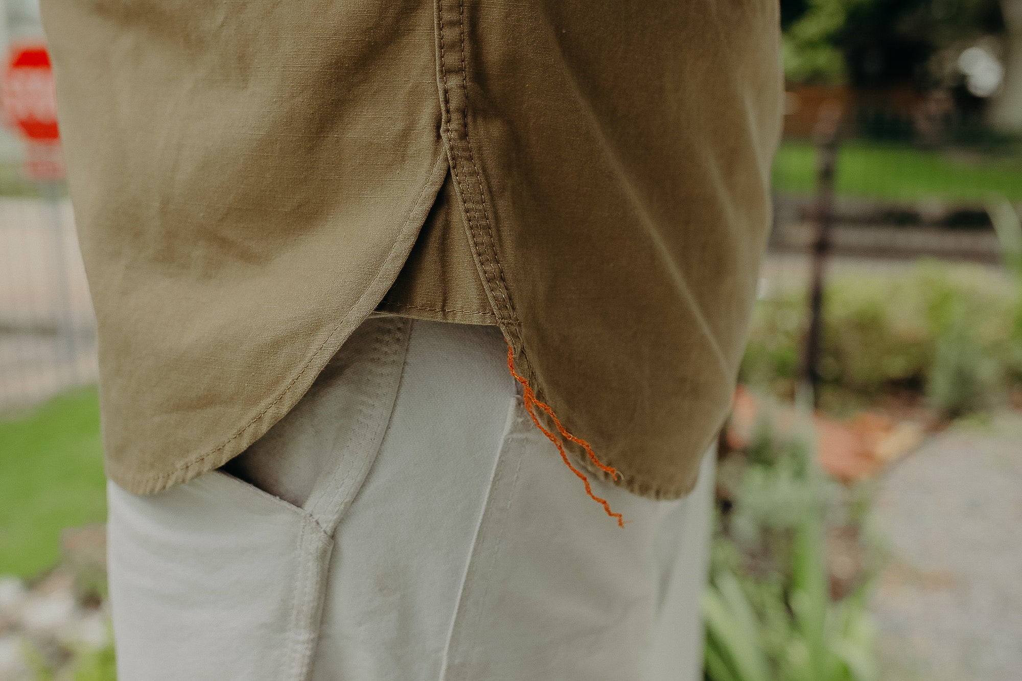 Close-up of brown shirt hem with orange thread detail over white pants in outdoor garden setting