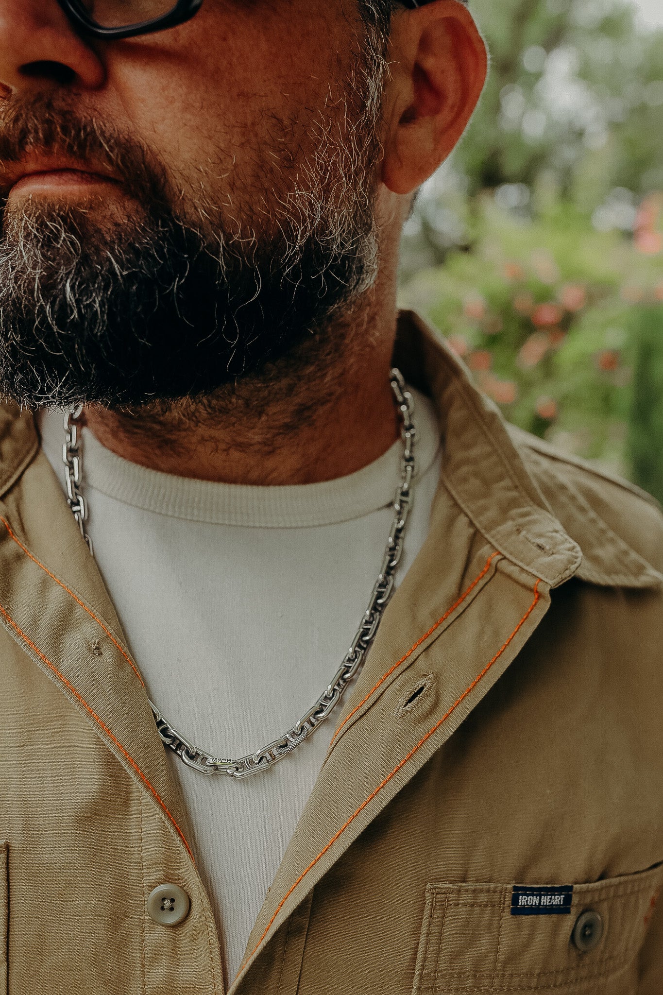 Close-up of a bearded man wearing glasses, a tan Iron Heart jacket with orange stitching, white shirt, and silver chain necklace