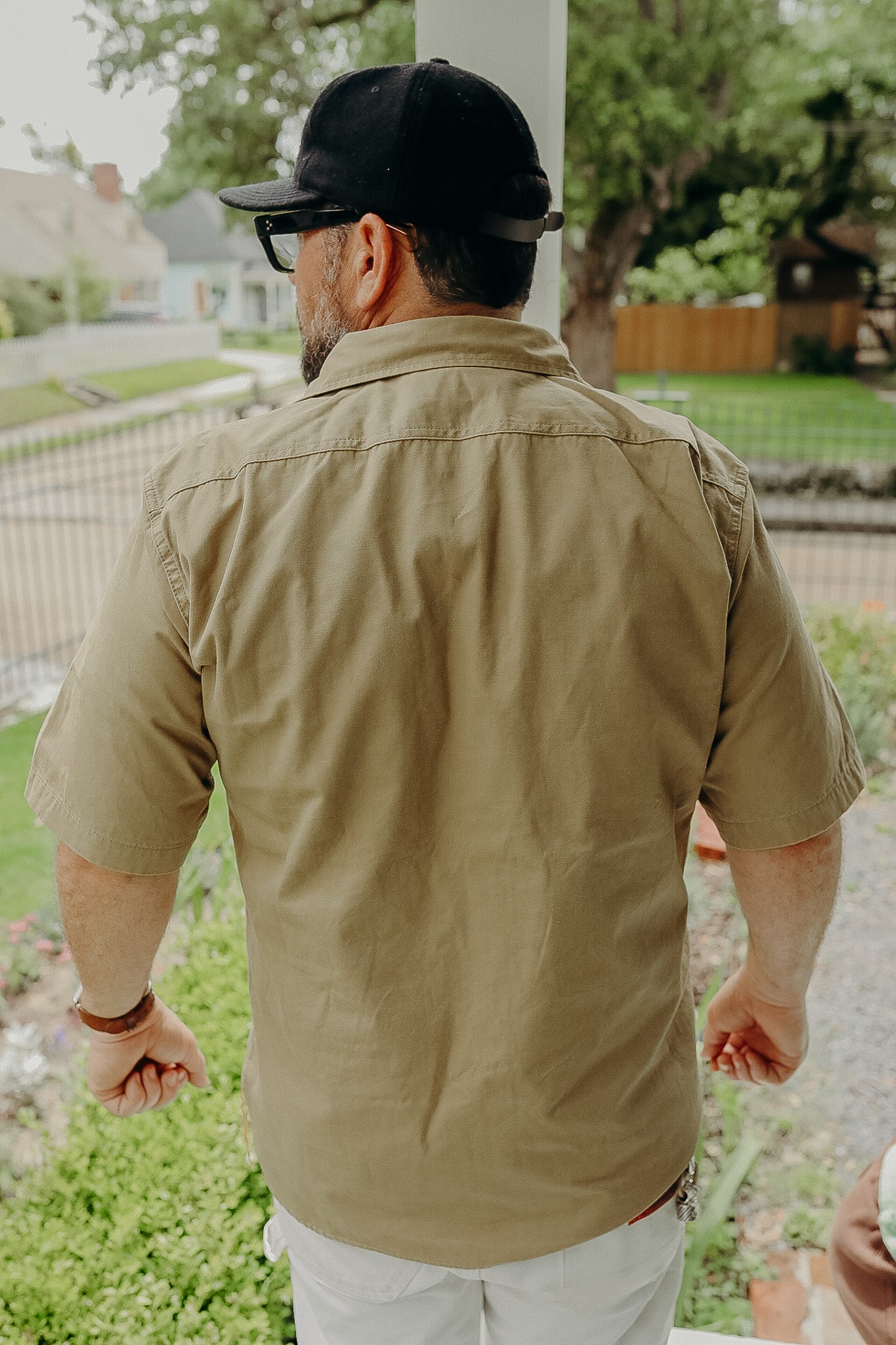 Man wearing khaki short-sleeve work shirt and black cap, standing outdoors on porch with garden background