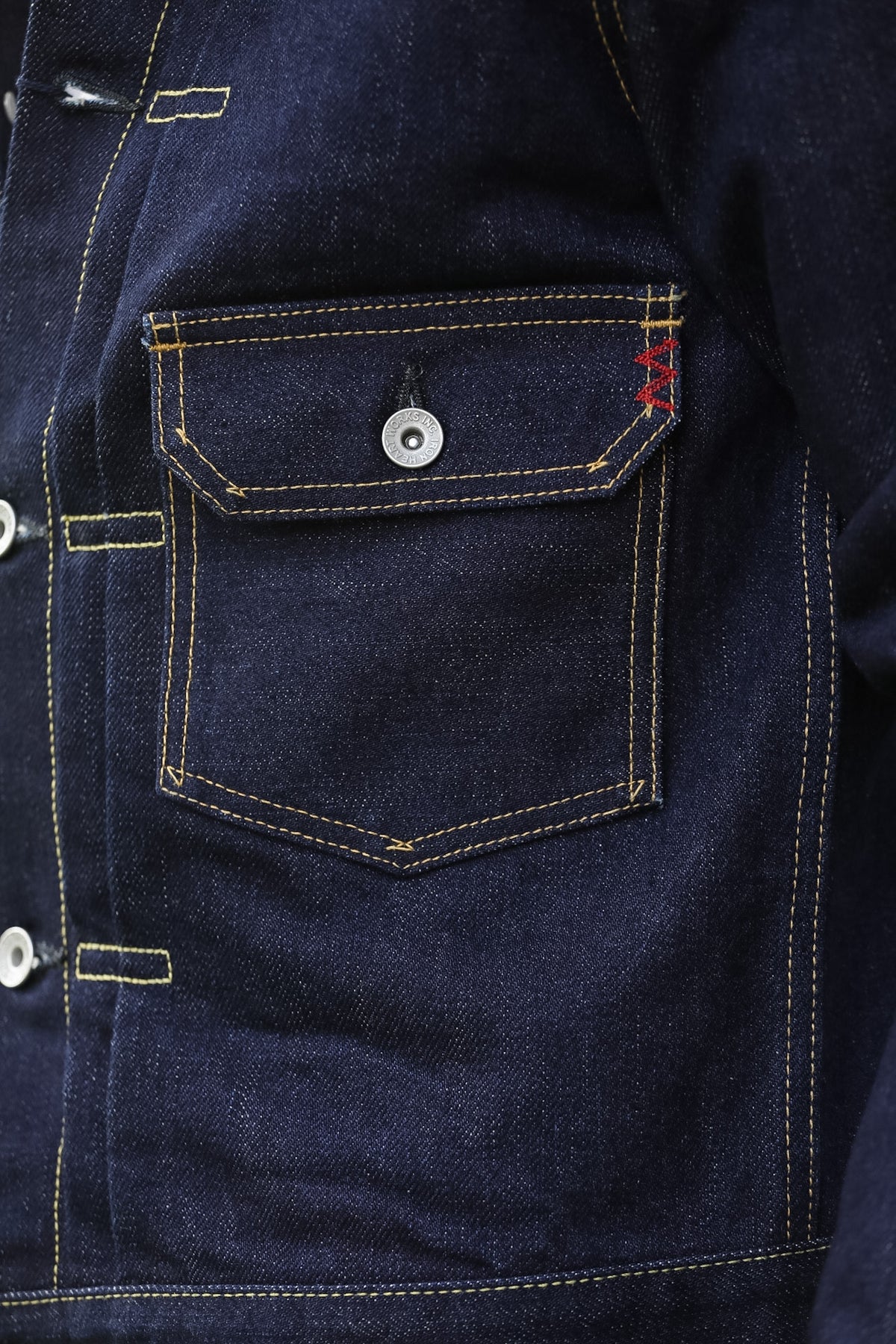 Close-up of a dark indigo denim jacket showing a front chest pocket with contrast yellow stitching, a silver snap button, and a small red embroidered logo on the pocket.