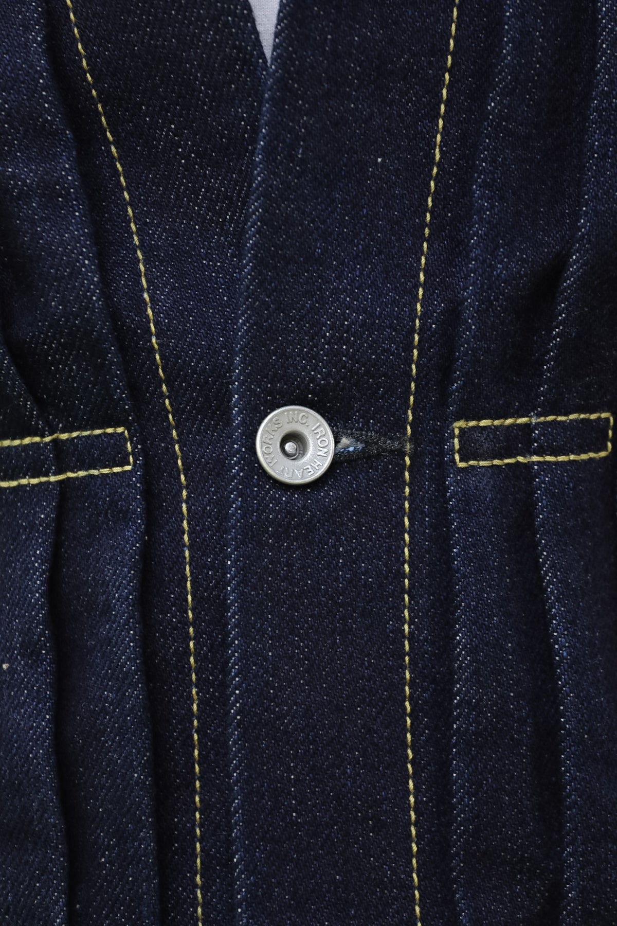 Close-up of a dark indigo denim garment, likely a jacket or overshirt, featuring yellow contrast stitching, a single silver metal snap button, and welt pockets.