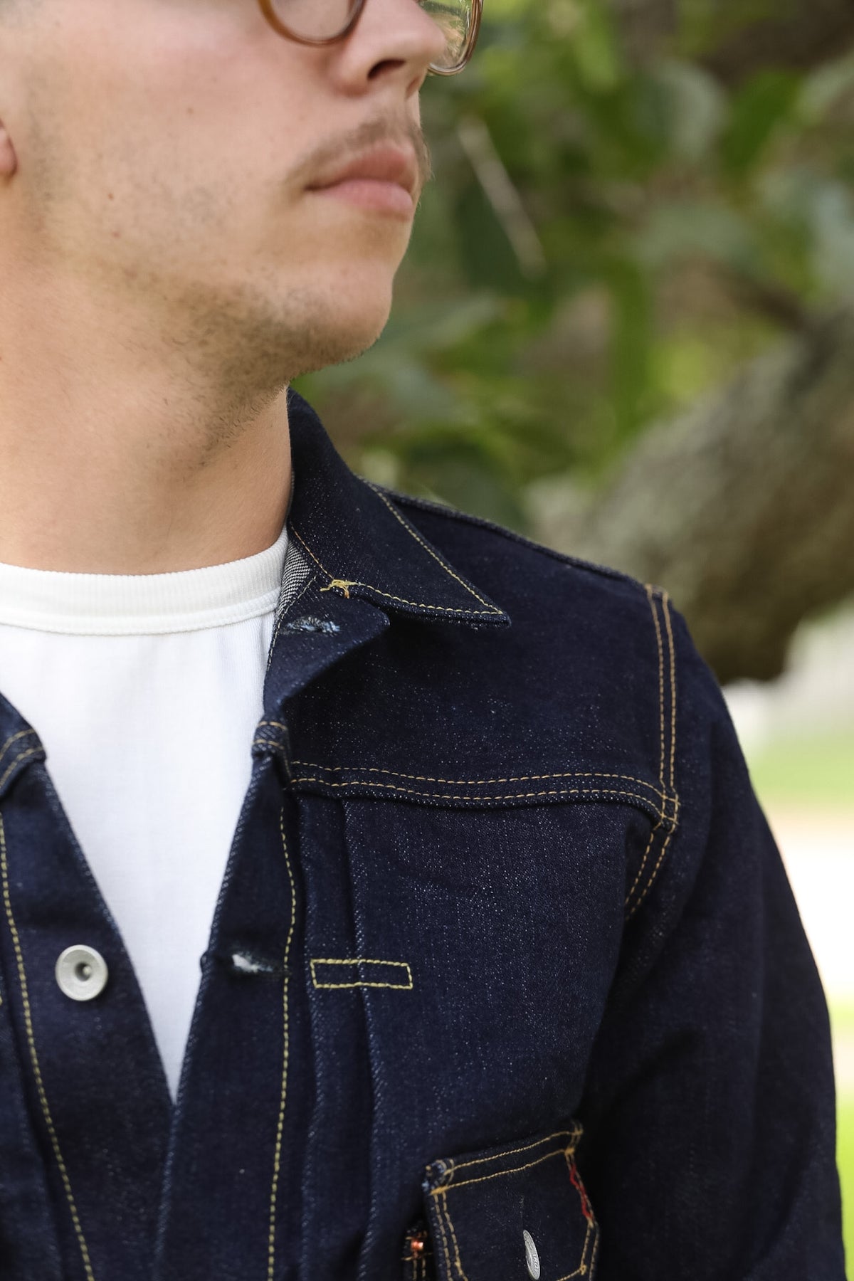 Close-up of a man wearing a dark indigo denim jacket over a white t-shirt, with a blurred outdoor background of green foliage.