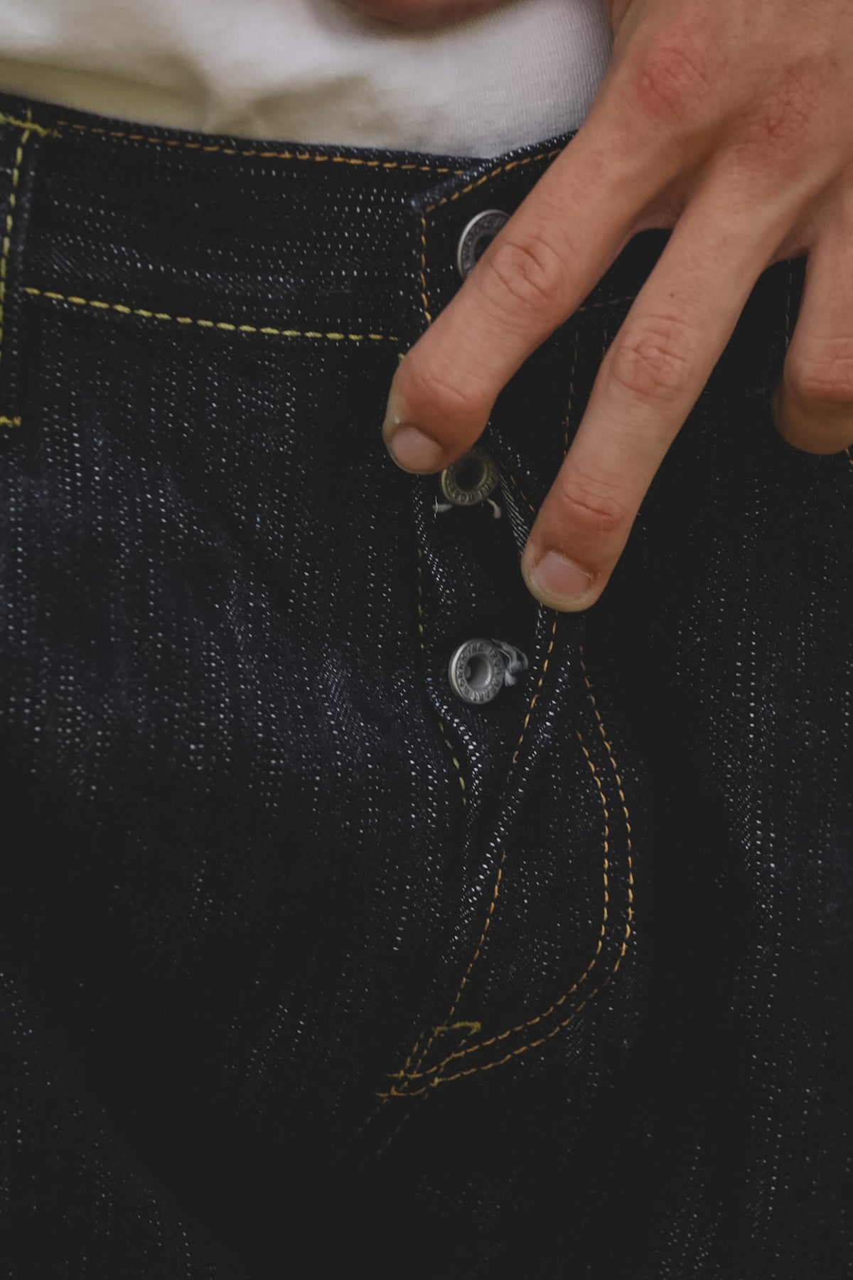 Close-up of a person's hand pulling a button on dark indigo denim jeans, showing the texture of the fabric and yellow contrast stitching.